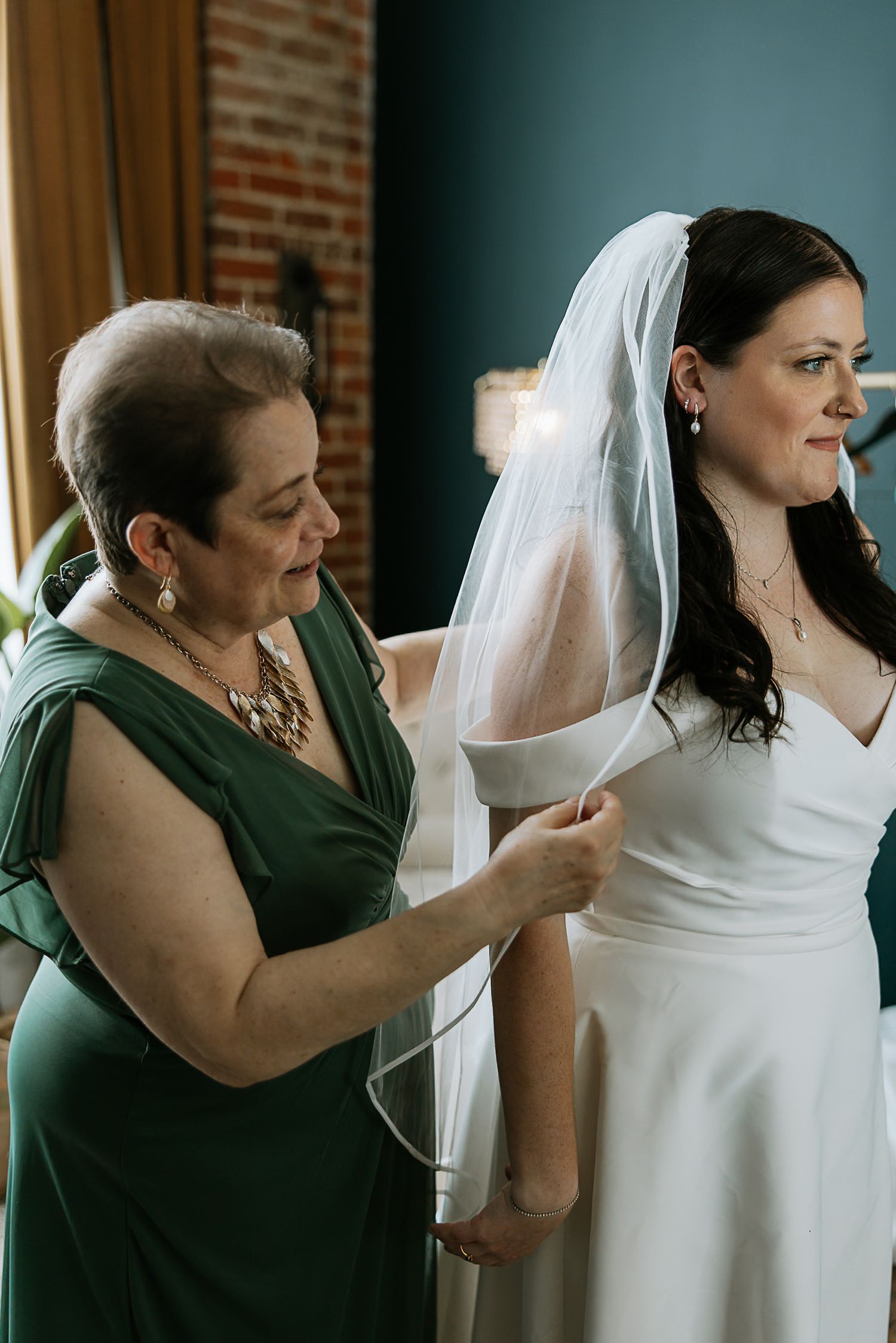 Mother in green dress carefully attaching veil to bride in white wedding gown.