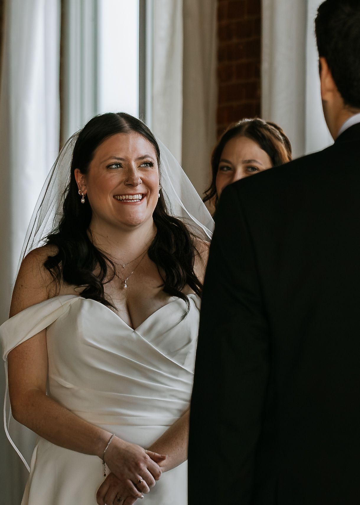 Radiant bride in white gown and veil smiling at groom during wedding ceremony.