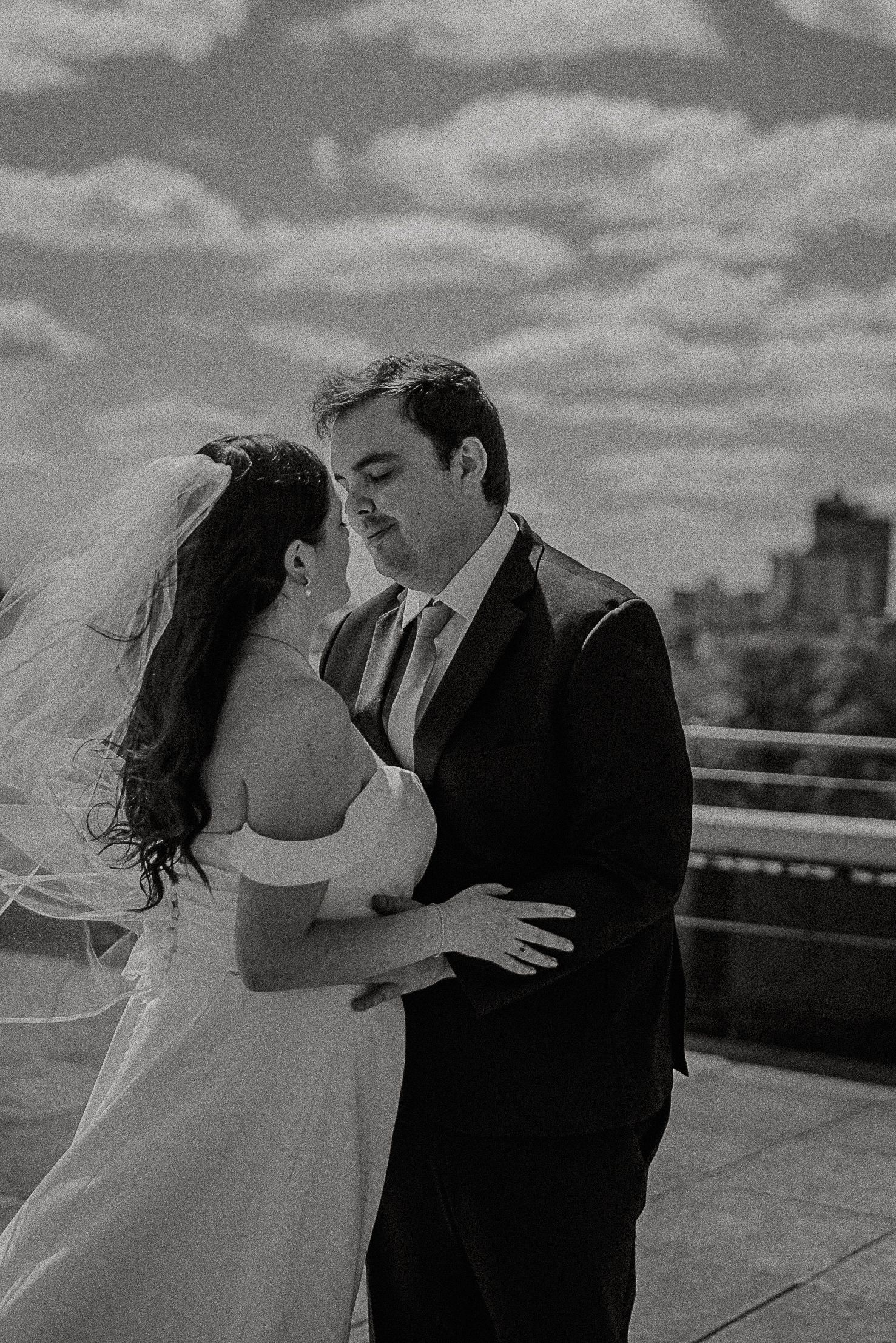 Black and white rooftop wedding photo of bride and groom embracing under dramatic cloudy sky.