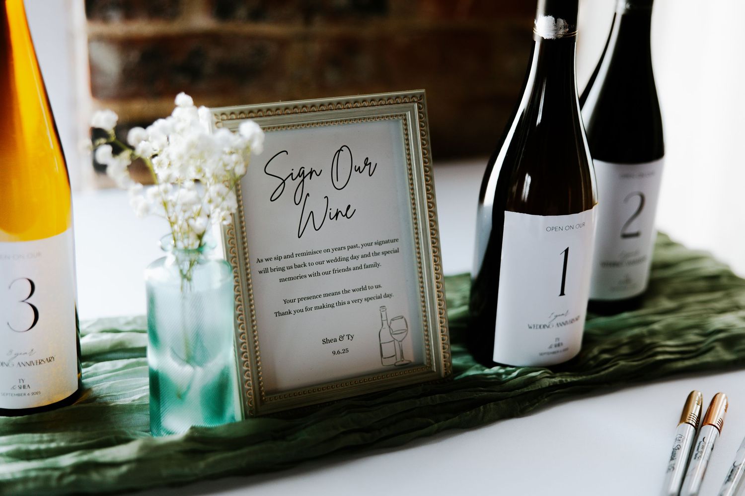 Wedding sign our wine station with framed sign, wine bottles, and paintbrushes on green velvet runner.