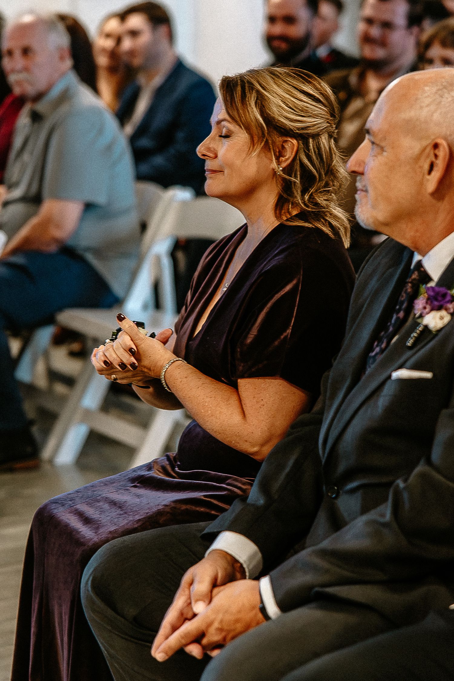 Emotional woman in dark velvet dress seated with man in suit watching wedding ceremony.