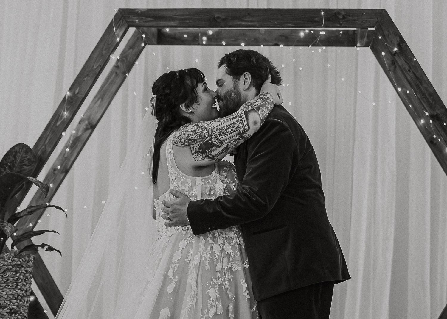 Black and white close-up of newlyweds sharing their first kiss under wooden arch.