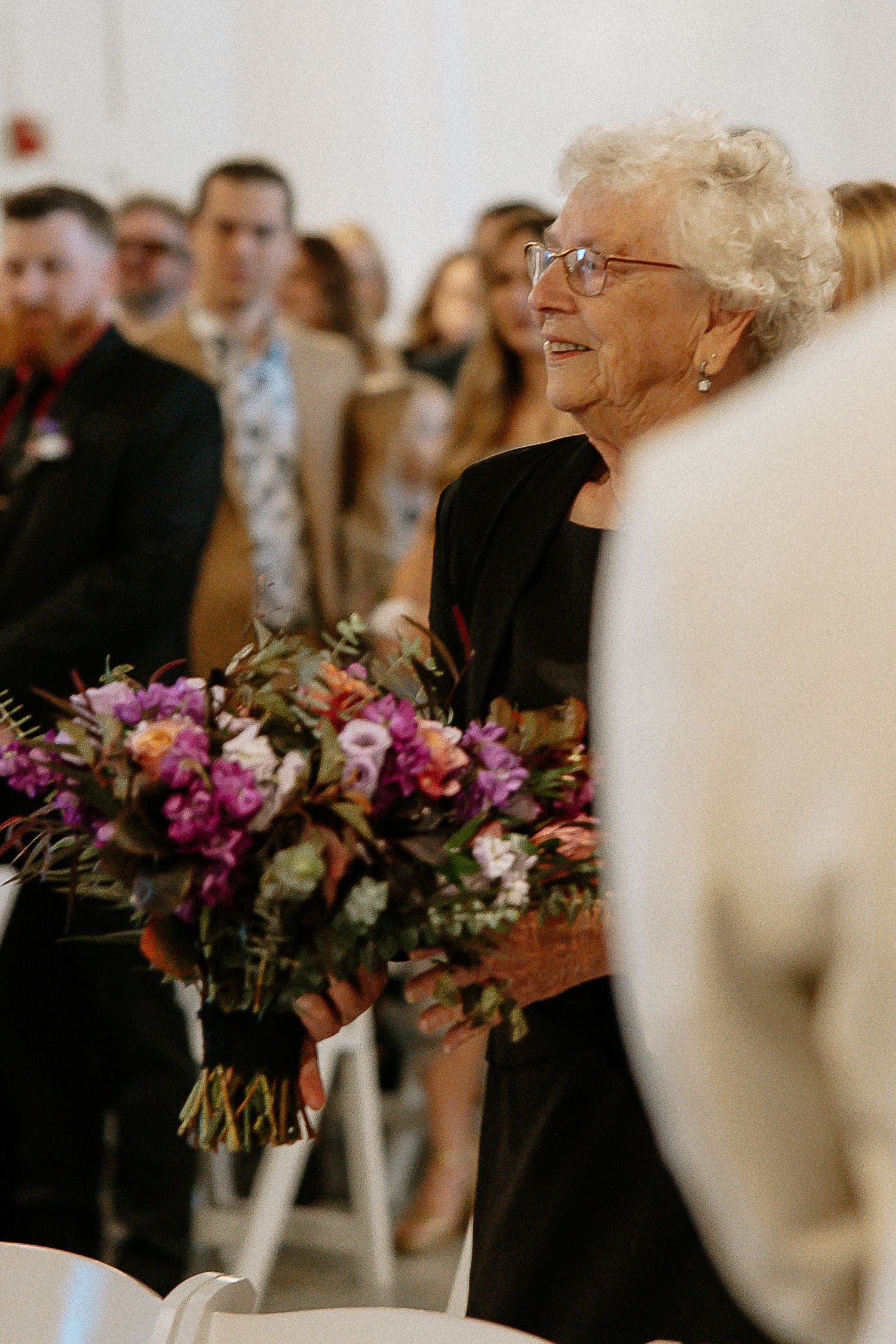 Elderly woman in black dress smiling while holding colorful bridal bouquets at a wedding ceremony.