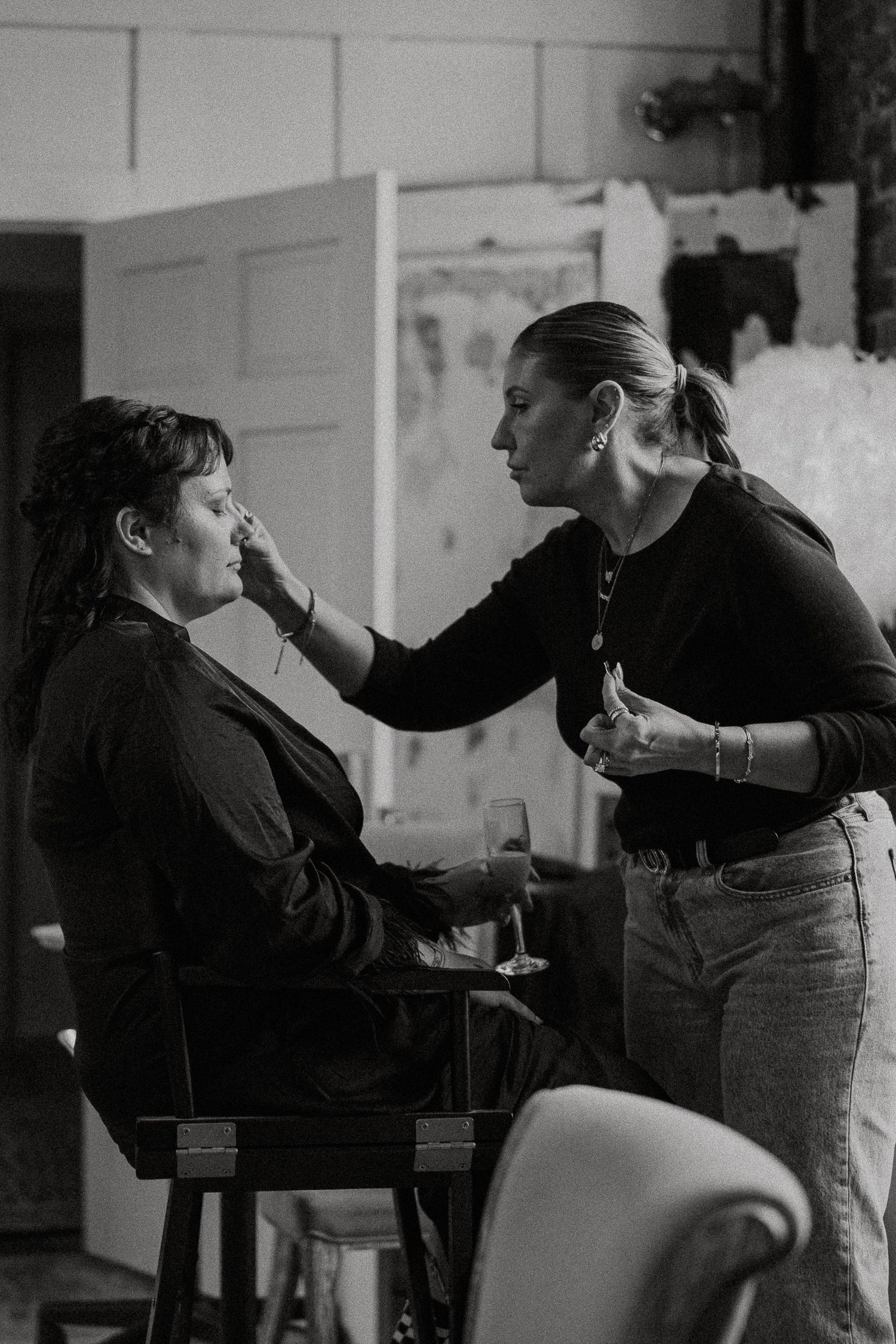 Makeup artist applies makeup to bride seated in chair during wedding prep.