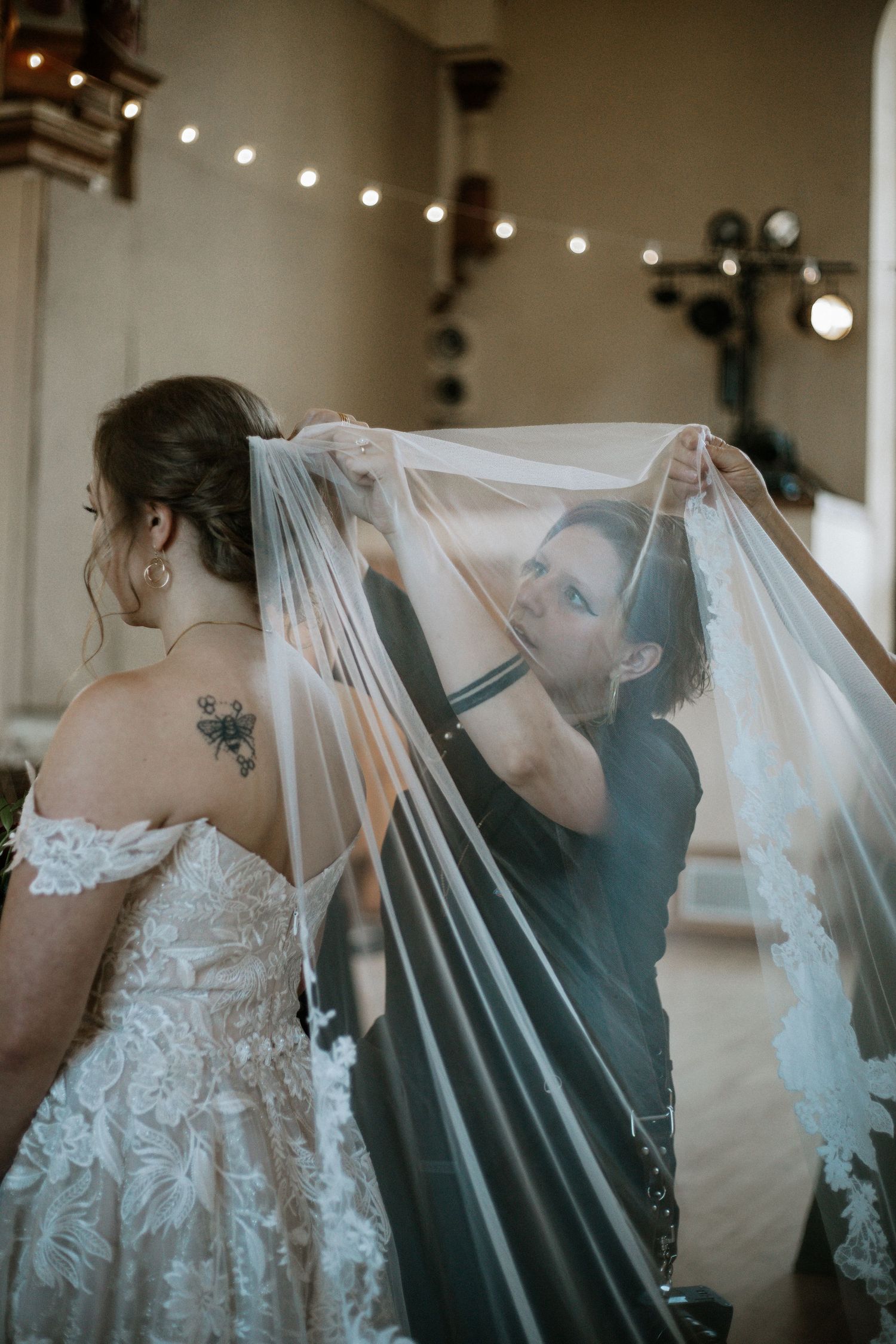 Wedding photographer adjusting brides long veil inside elegant venue with string lights.