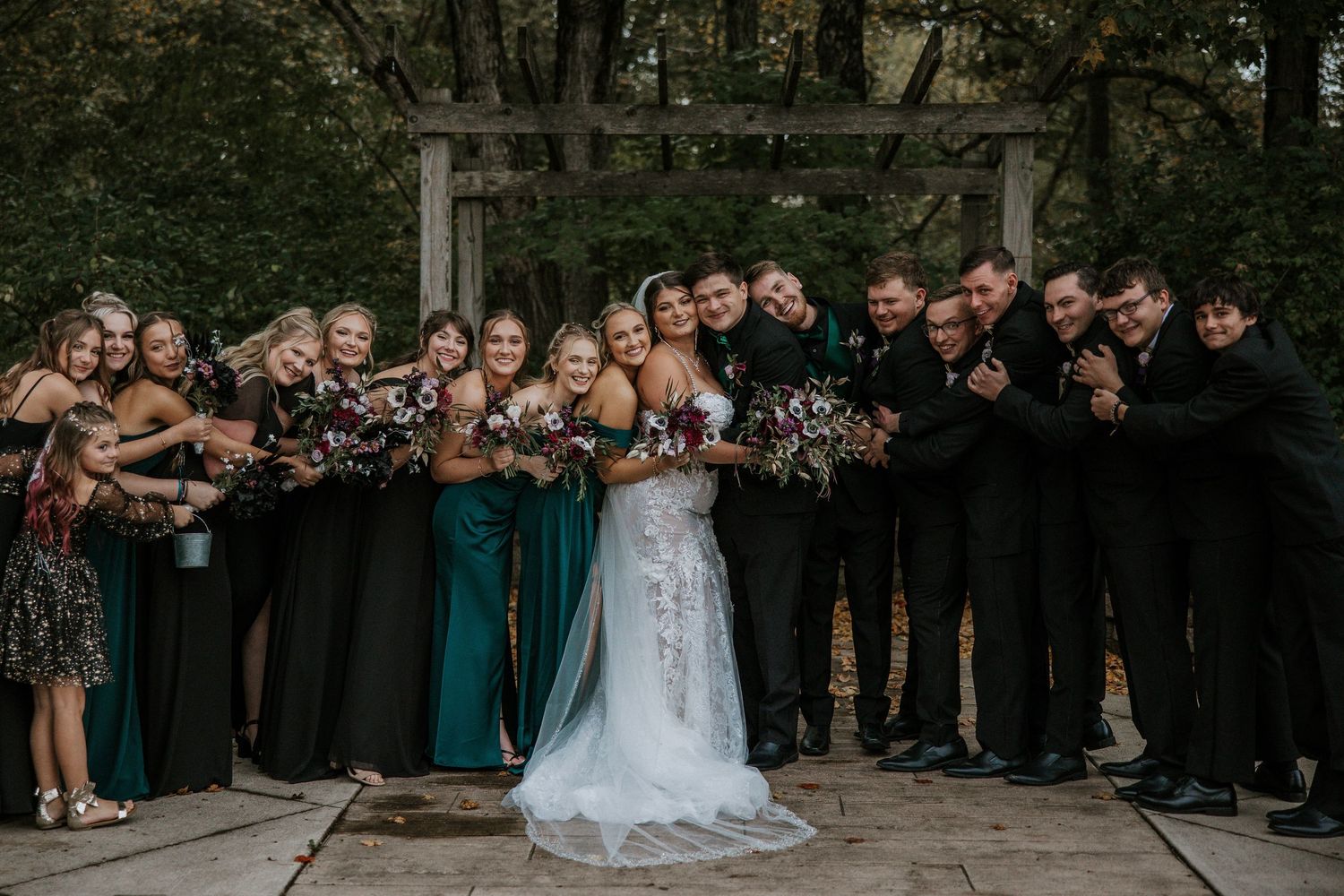 Wedding party poses together outdoors with bridesmaids in emerald green dresses and groomsmen in black suits.