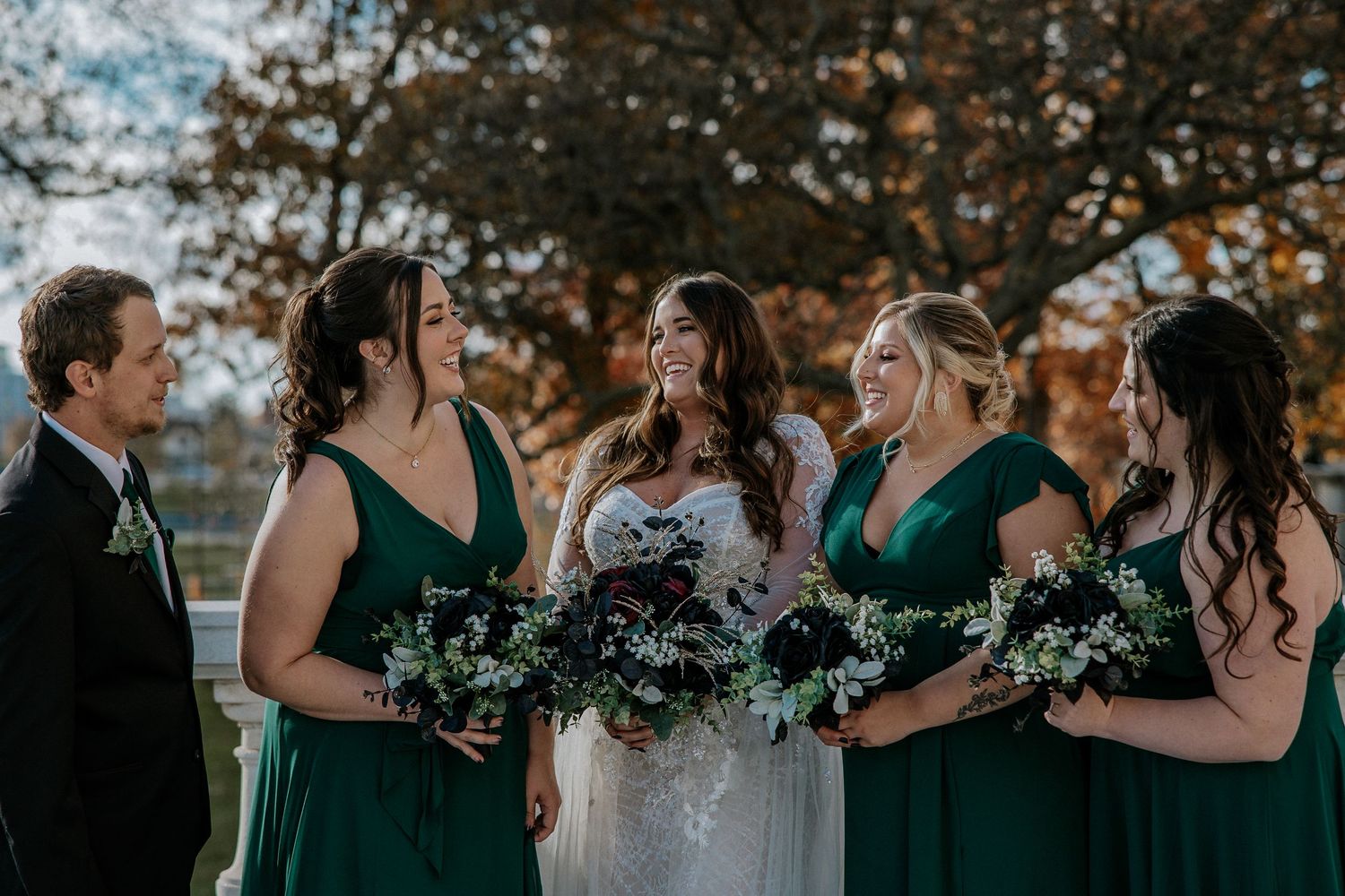 Bride surrounded by bridesmaids in emerald green dresses holding bouquets of dark florals outdoors.