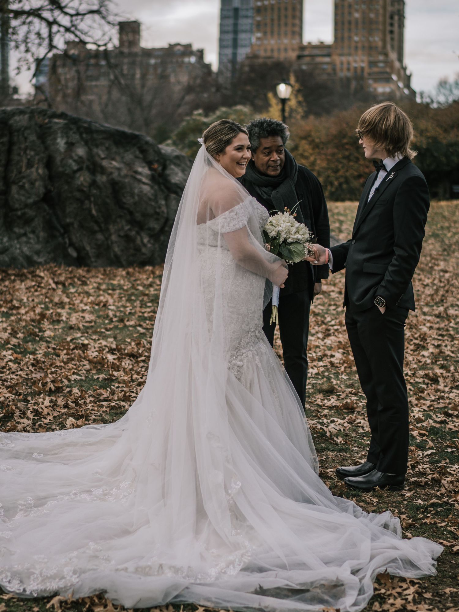 Bride in elegant mermaid wedding dress with long veil stands with groom and officiant in Central Park fall foliage.