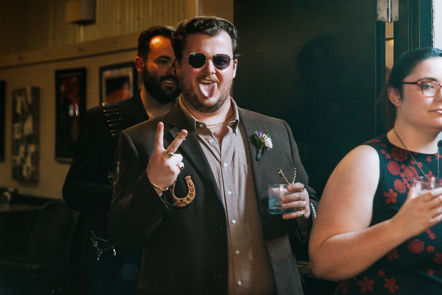 Man in sunglasses making rock sign at wedding reception in dark suit with boutonniere.