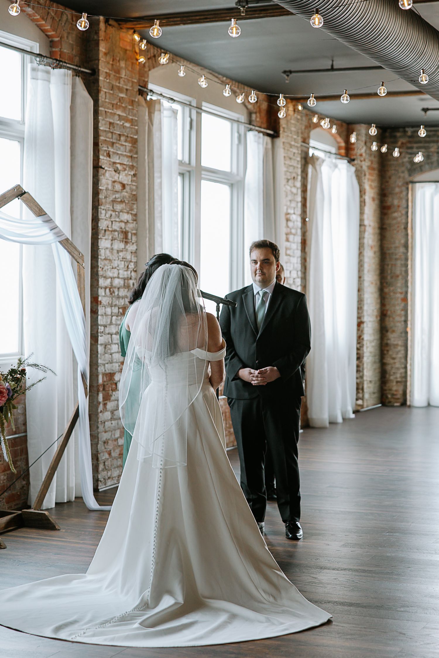Bride in white gown with veil faces groom during indoor wedding ceremony at rustic venue.