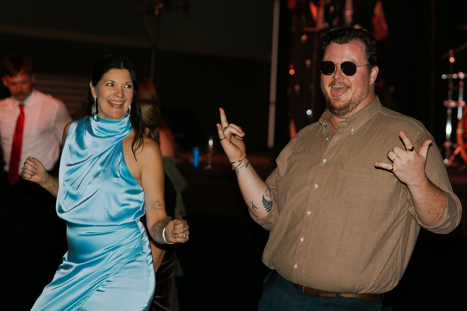Couple dances joyfully at wedding reception, woman in blue satin dress, man in tan shirt.