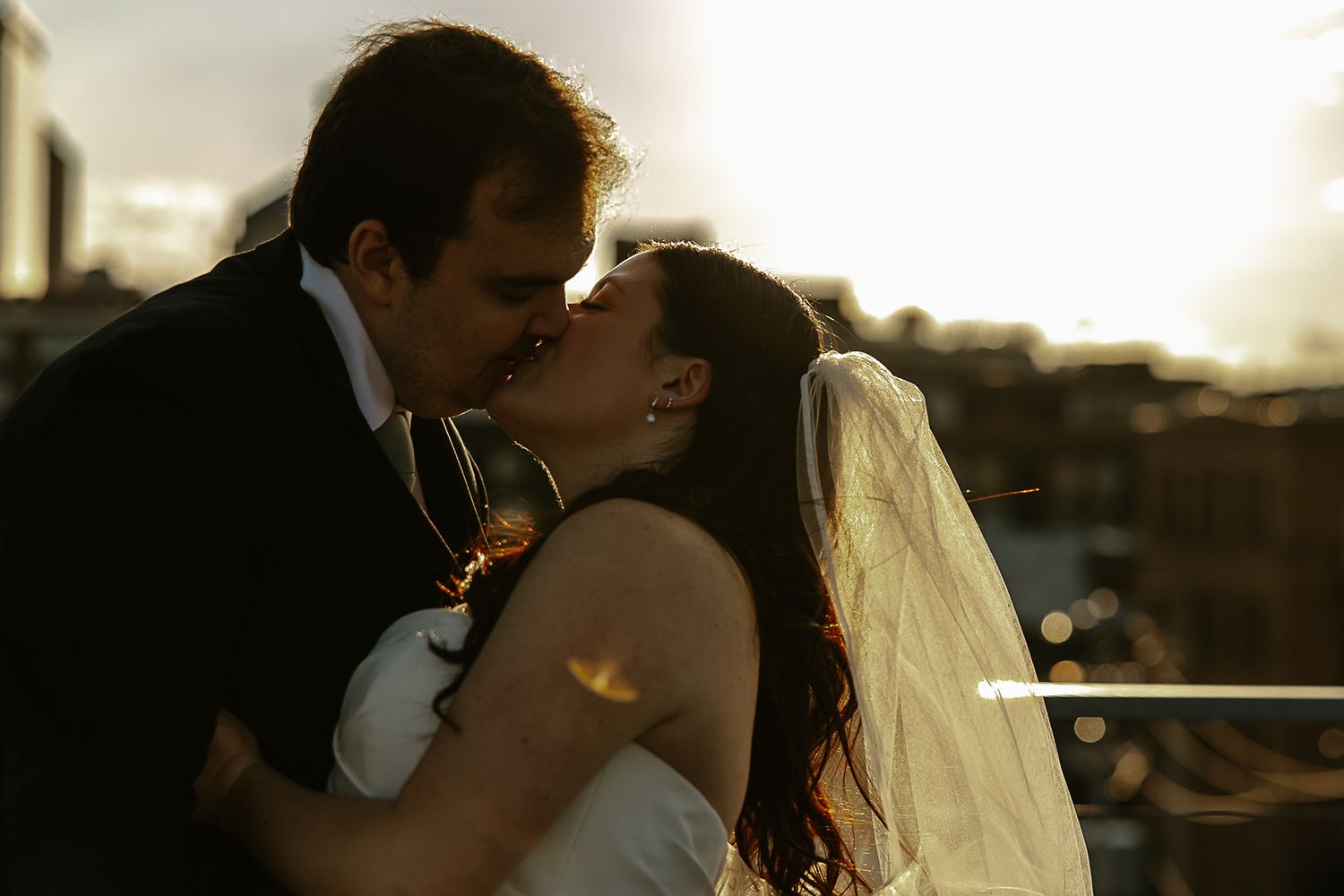 Bride and groom sharing a romantic kiss at sunset with city backdrop.
