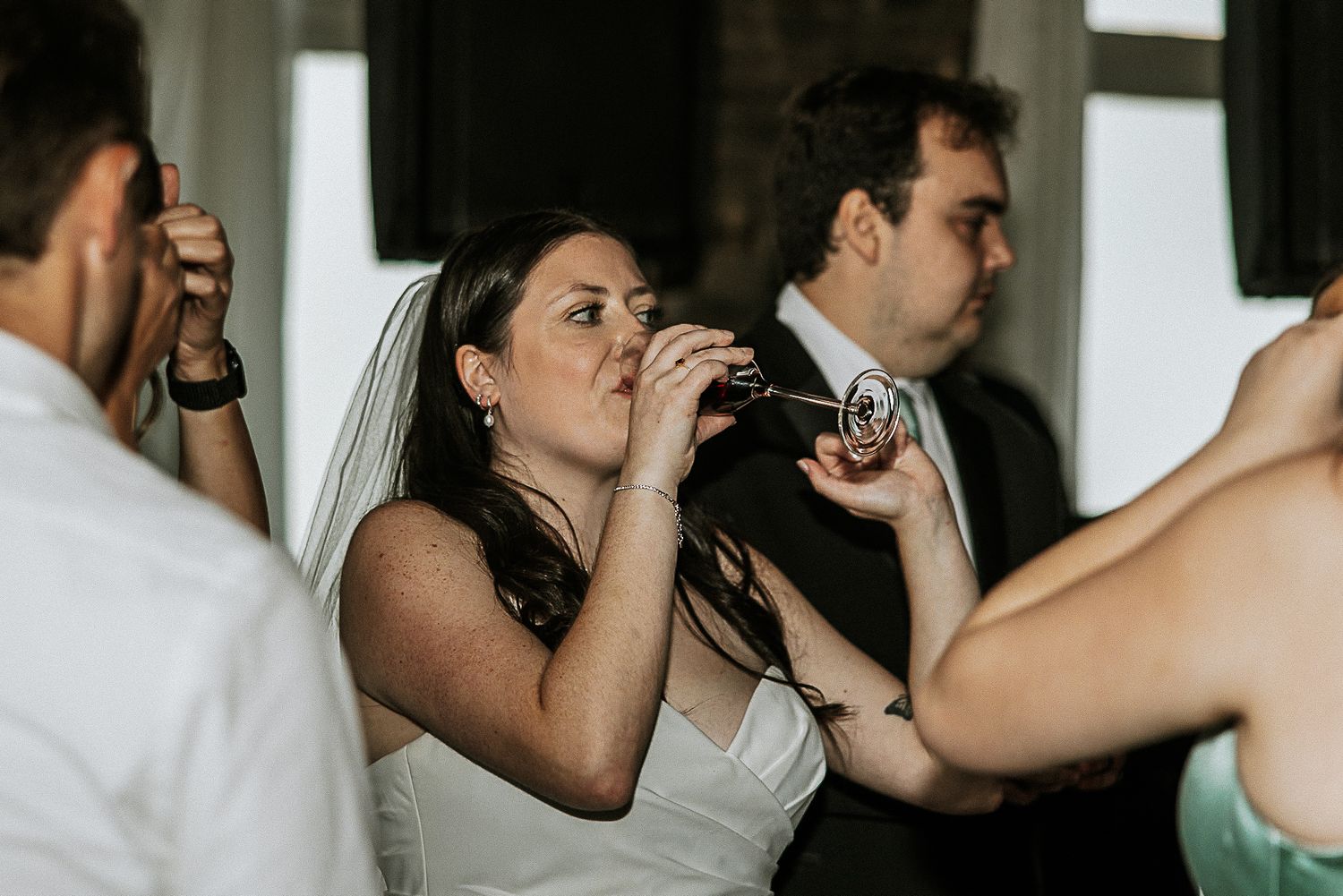 Bride in white wedding dress takes a celebratory shot drink during lively wedding reception party.