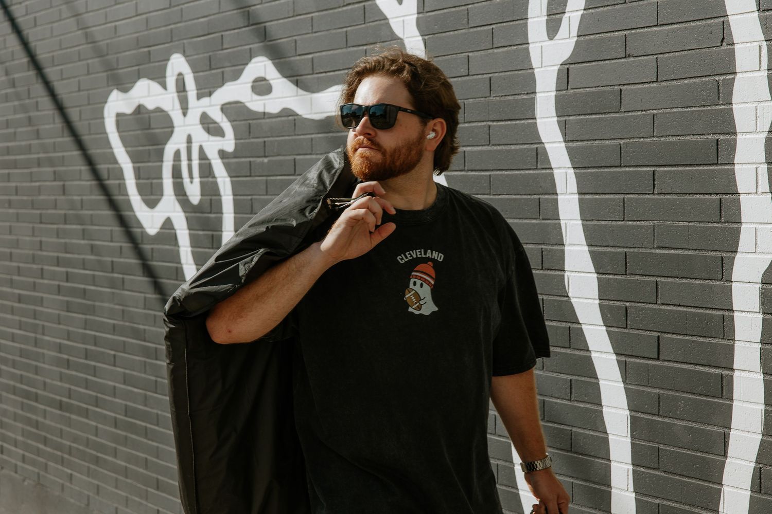 Bearded man in black graphic tee wearing sunglasses posing against graffiti brick wall.