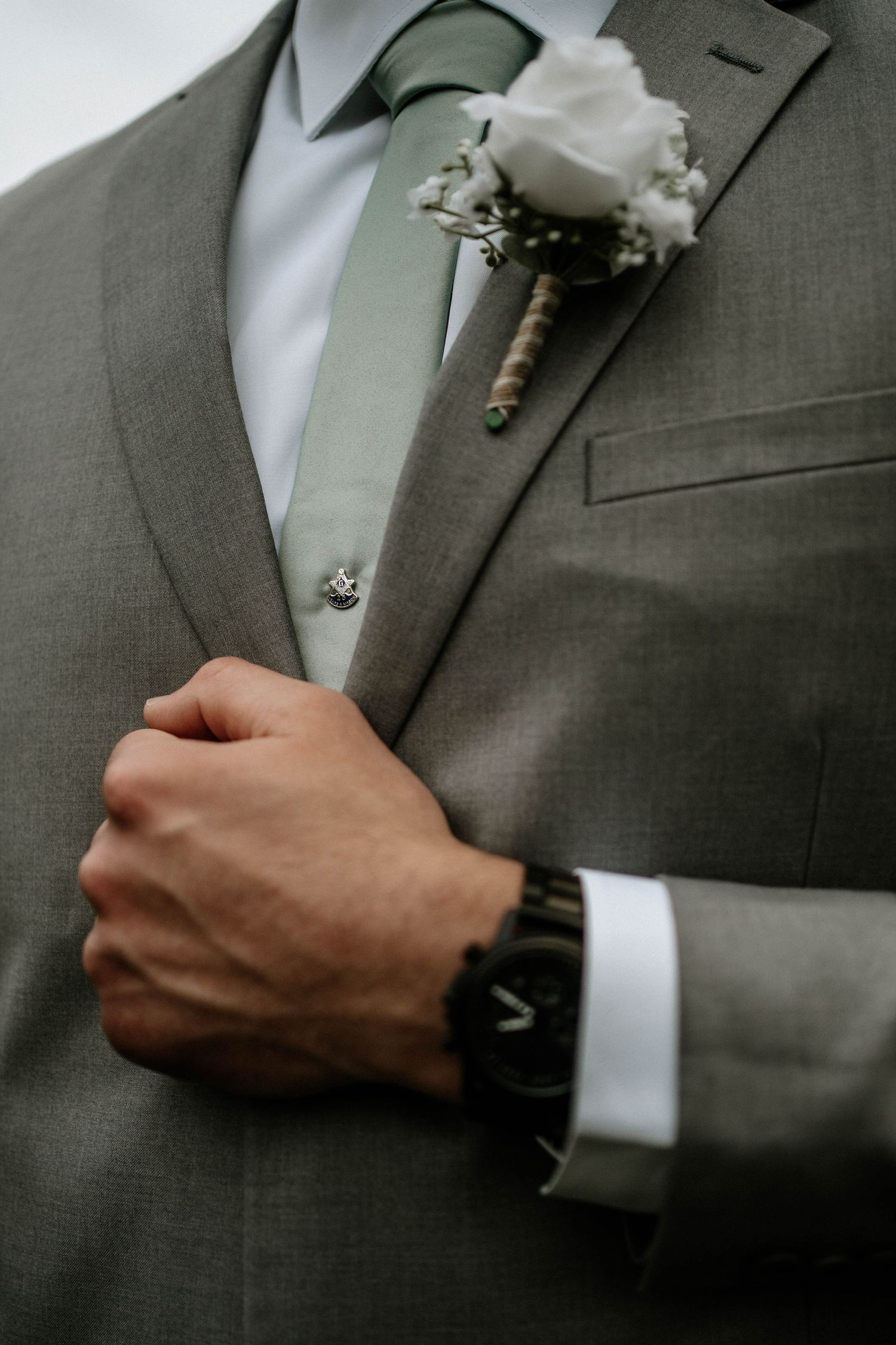 Groom in gray suit with sage green tie and white rose boutonniere adjusting jacket lapel.