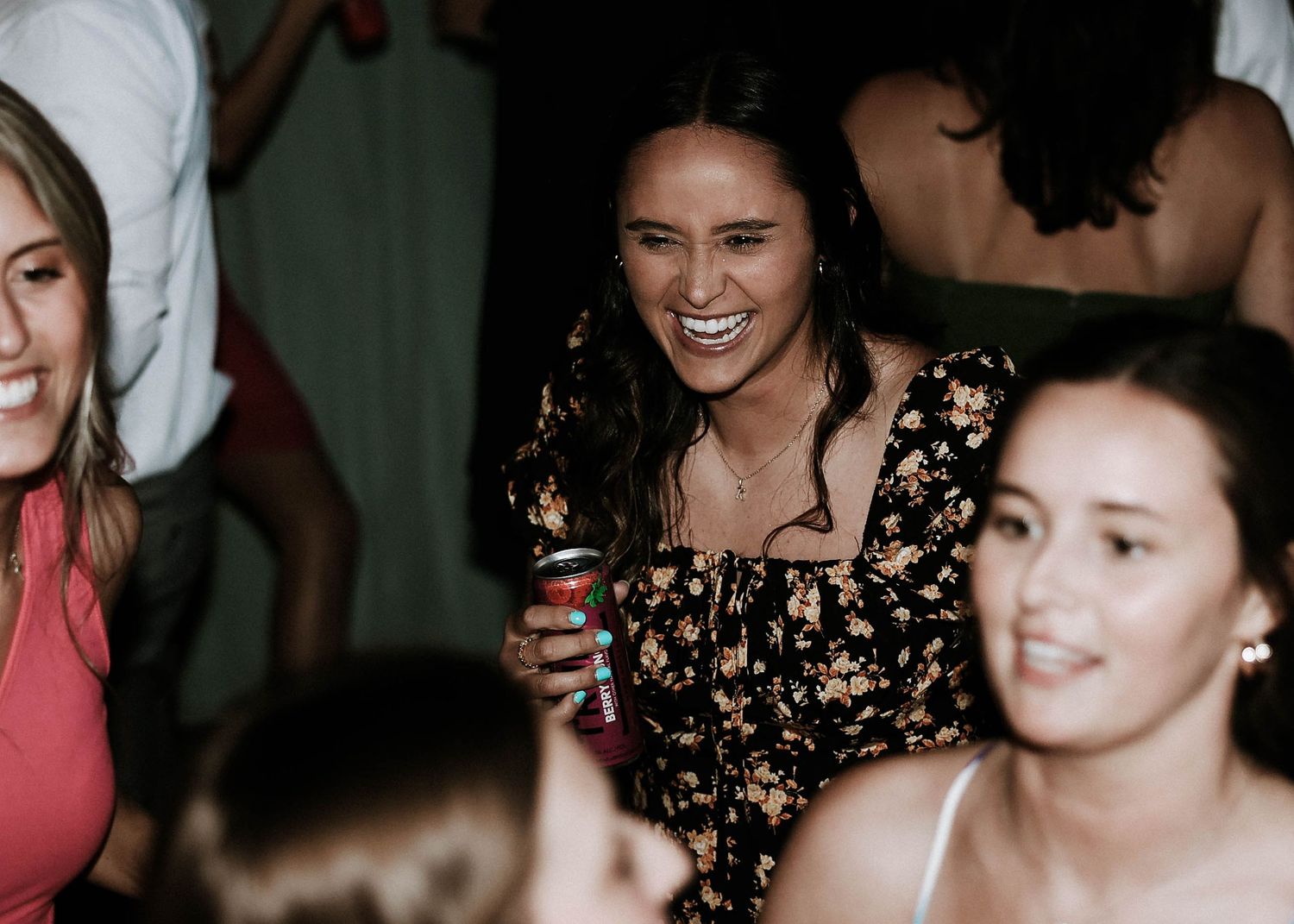 Woman in floral dress laughing and holding a drink at a lively party with friends.