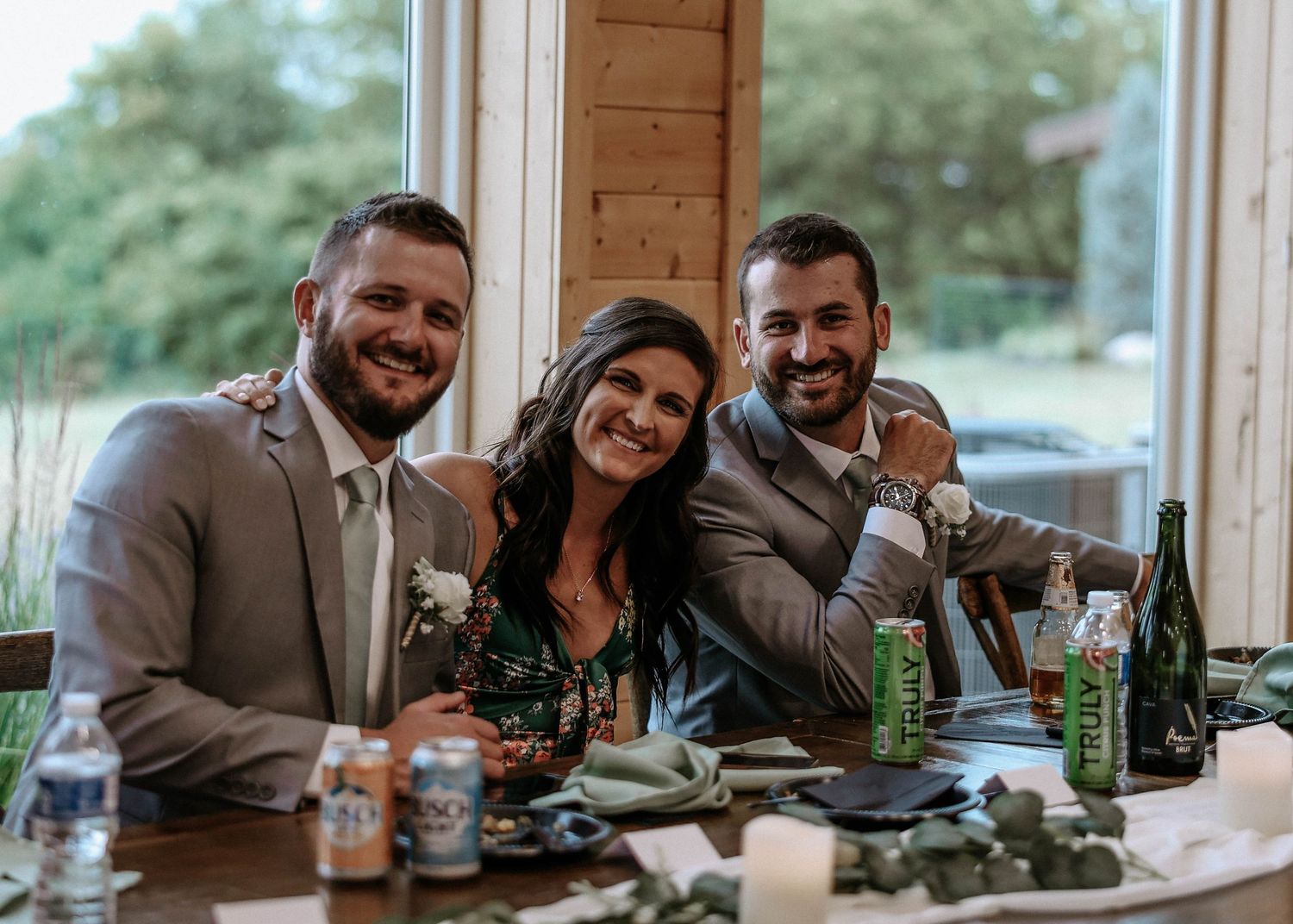 Three smiling wedding guests pose together at a decorated reception table with drinks and greenery.