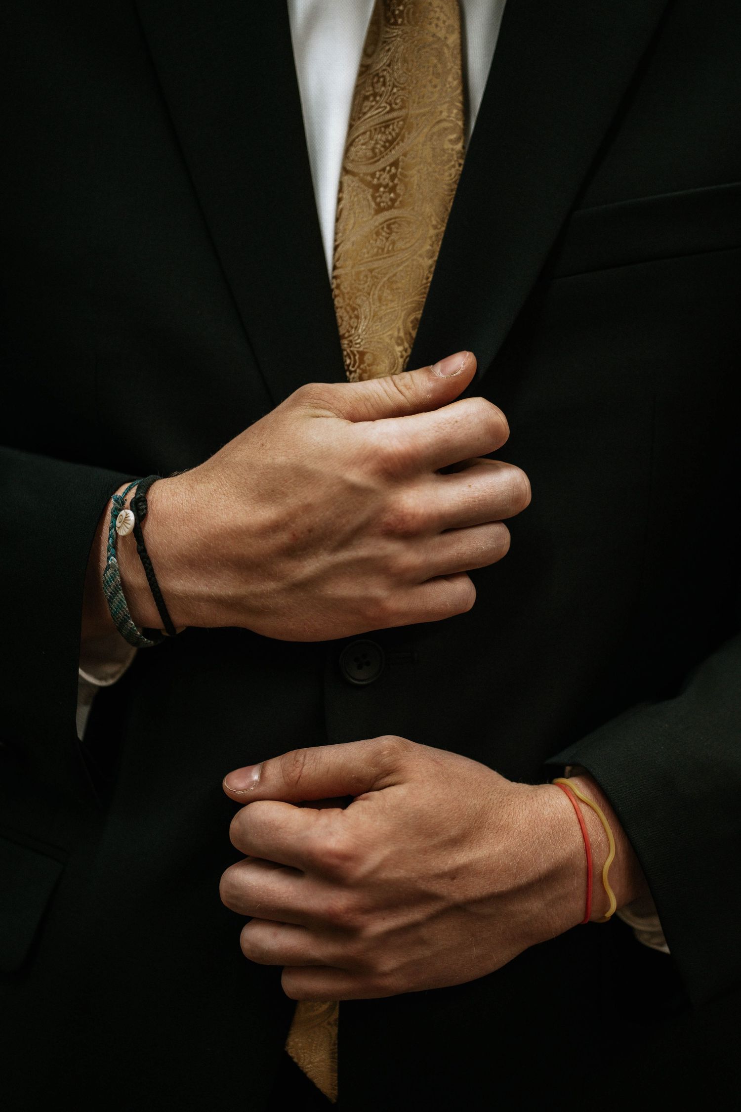 Man in black suit adjusting jacket with gold paisley tie, wearing bracelet and red string bracelet.