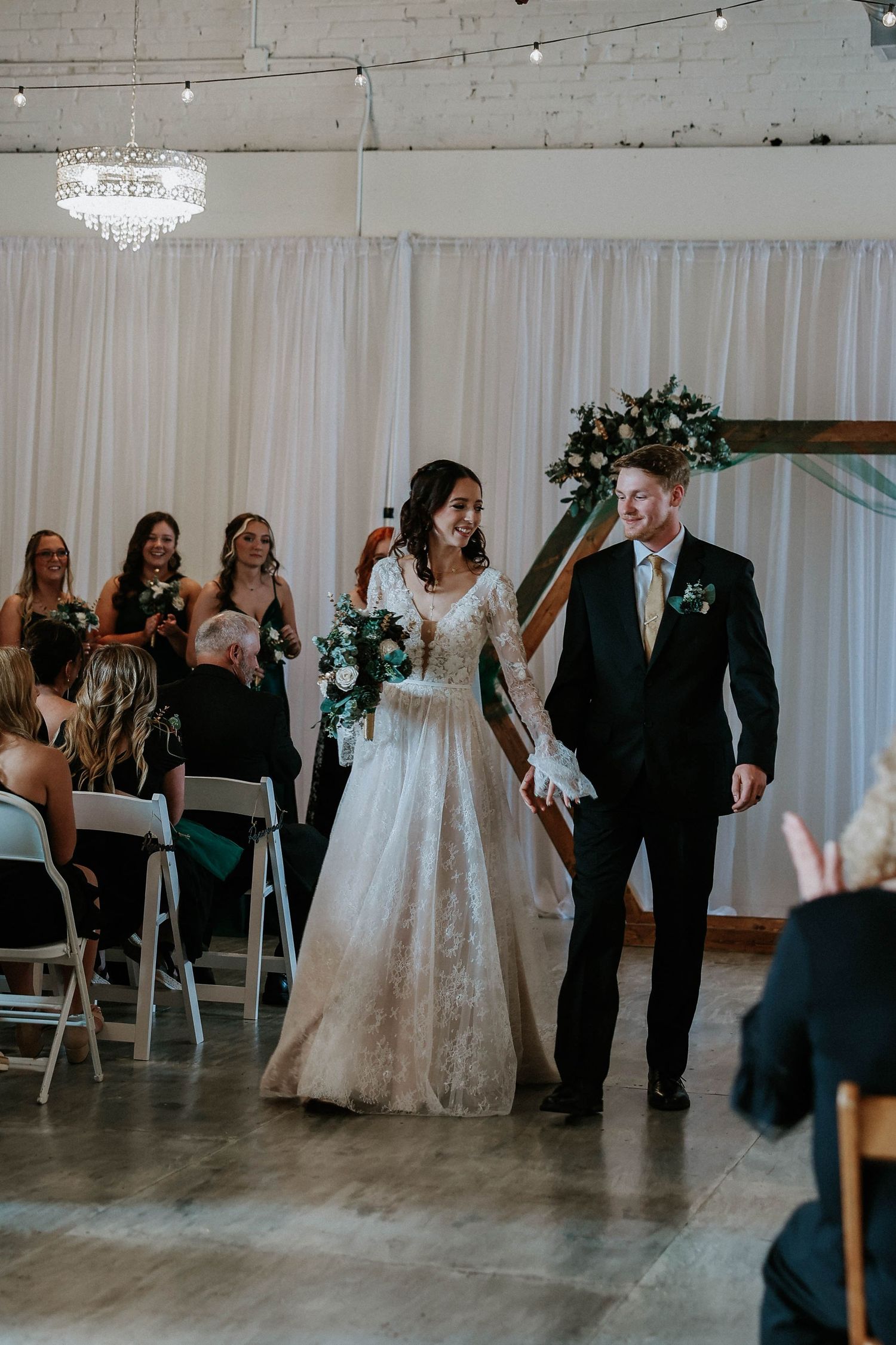 Bride in lace gown and groom in black tuxedo walk down the aisle smiling at their indoor wedding ceremony.