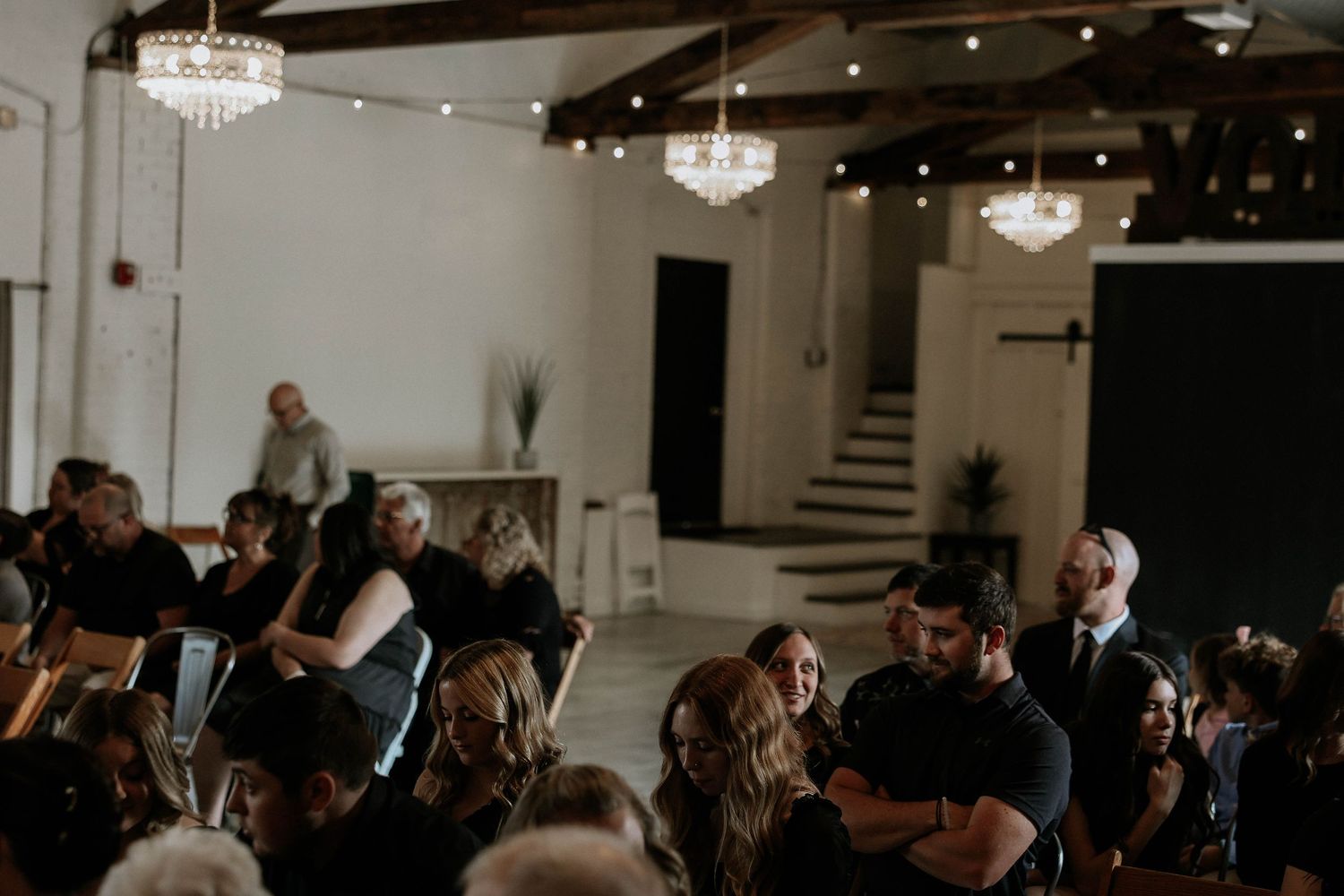 Elegant wedding ceremony venue with chandeliers, string lights, and guests seated in rows on wooden chairs.