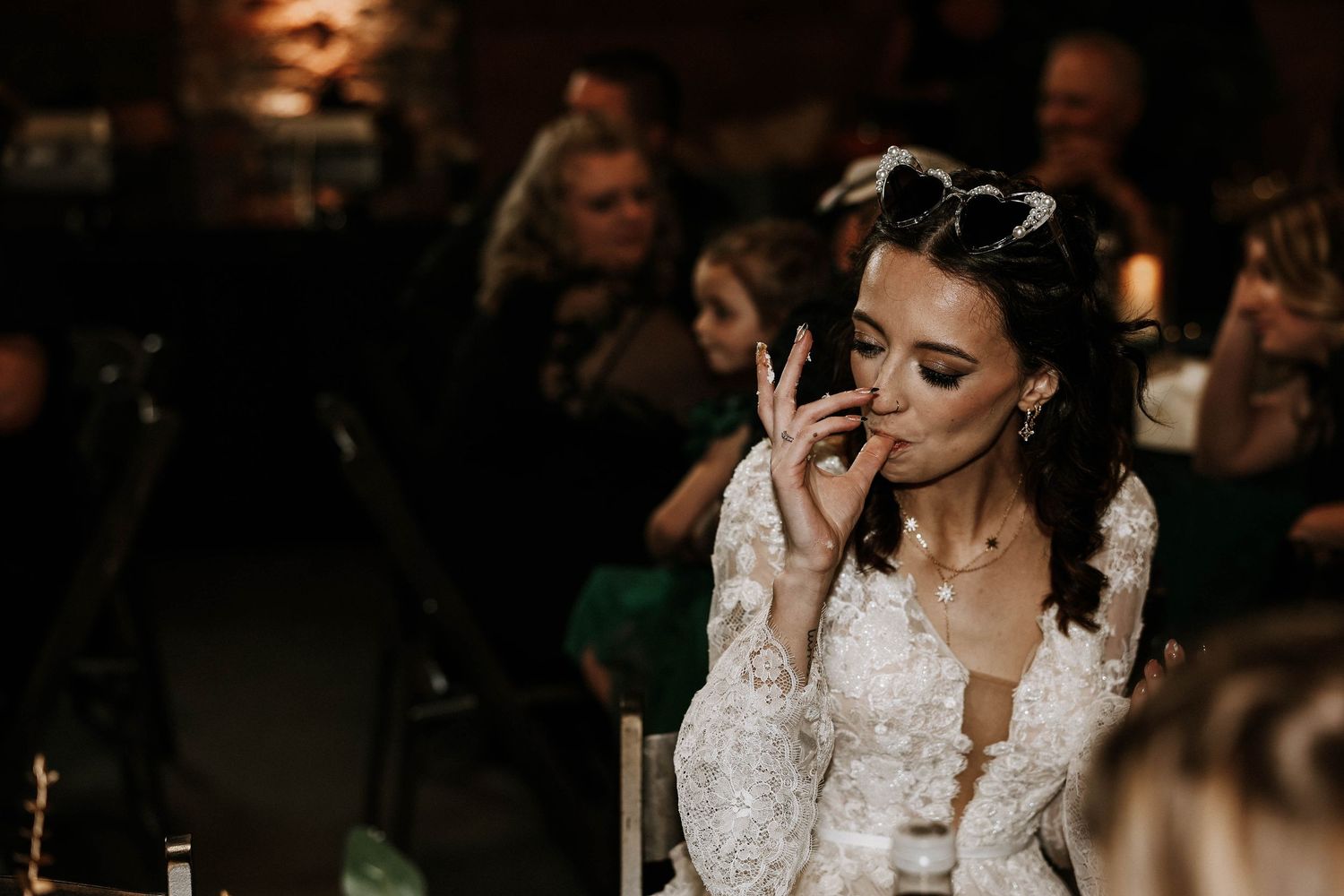 Bride in lace wedding dress kissing her hand at a dimly lit wedding reception with guests in background.