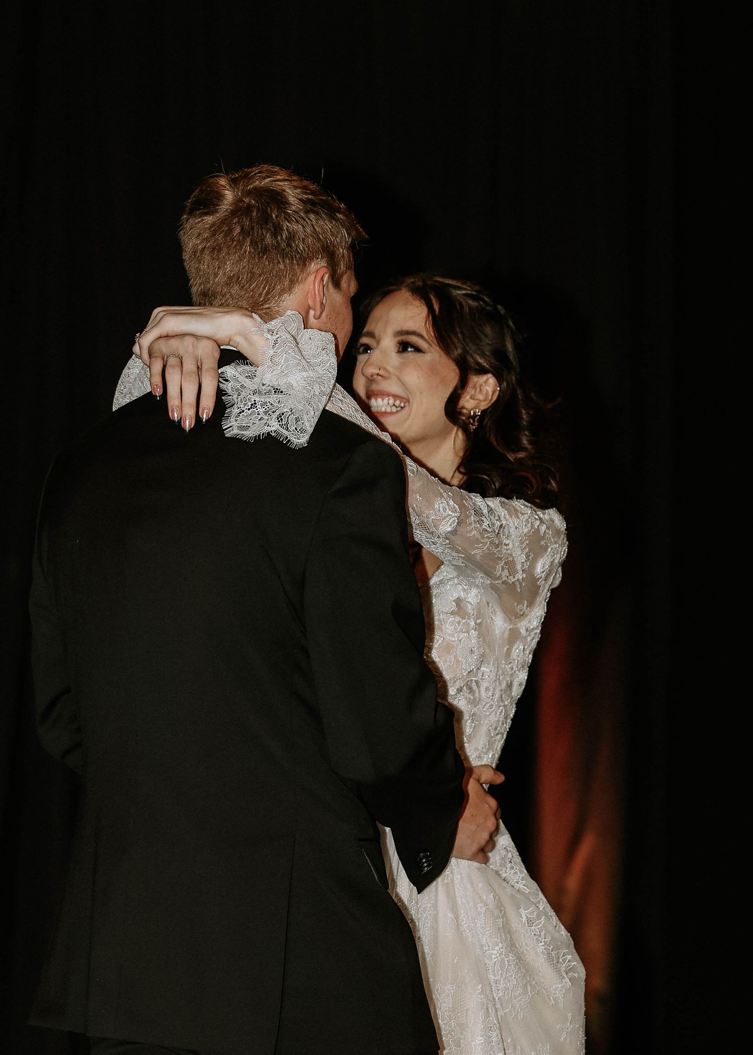 Bride in lace gown smiling joyfully during first dance with groom in black tuxedo at wedding reception.
