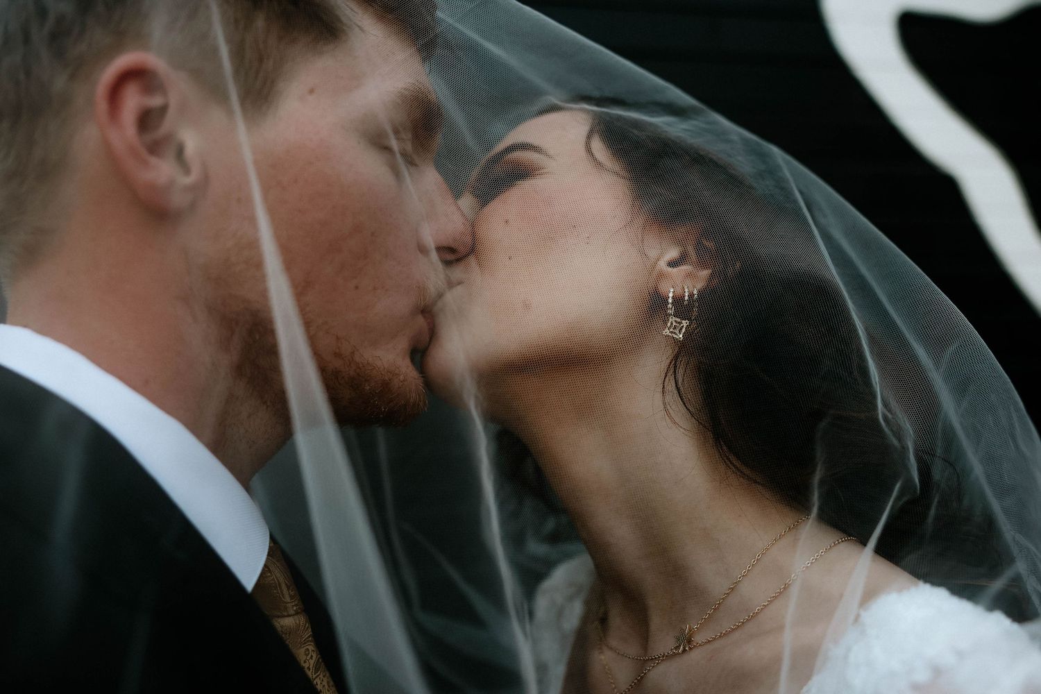 A bride and groom share a romantic kiss under a flowing white wedding veil during their outdoor ceremony.