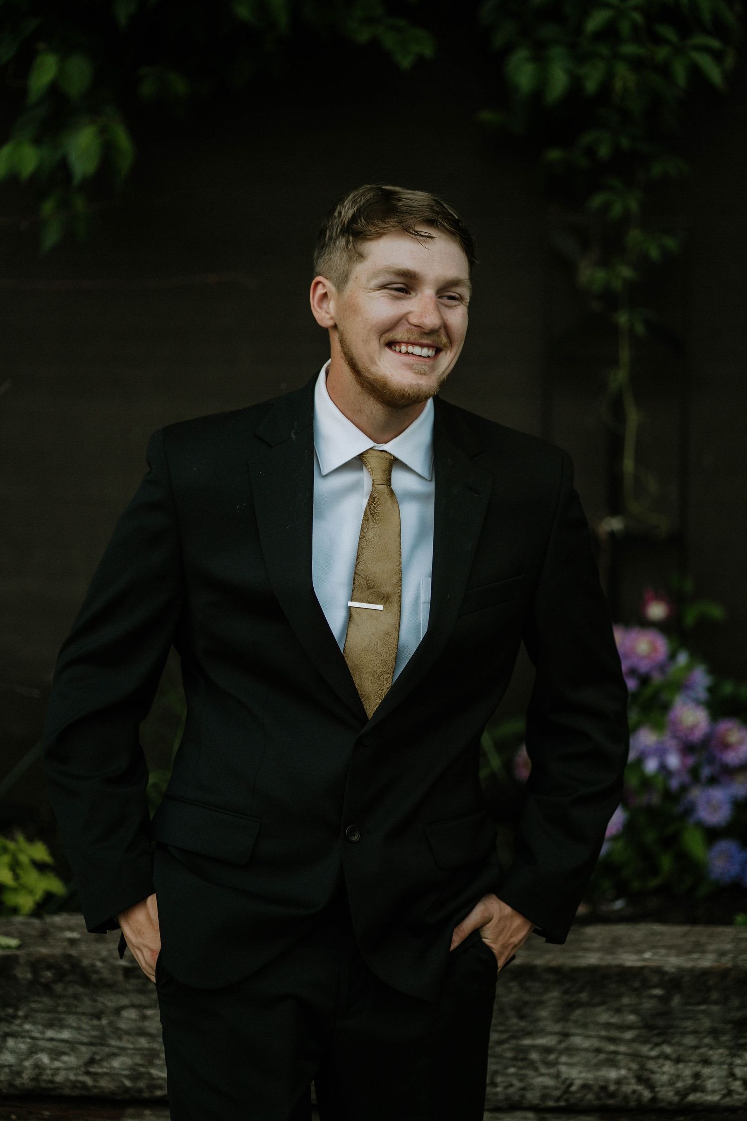 Smiling young man in black suit with gold tie and tie clip, standing outdoors with green foliage background.
