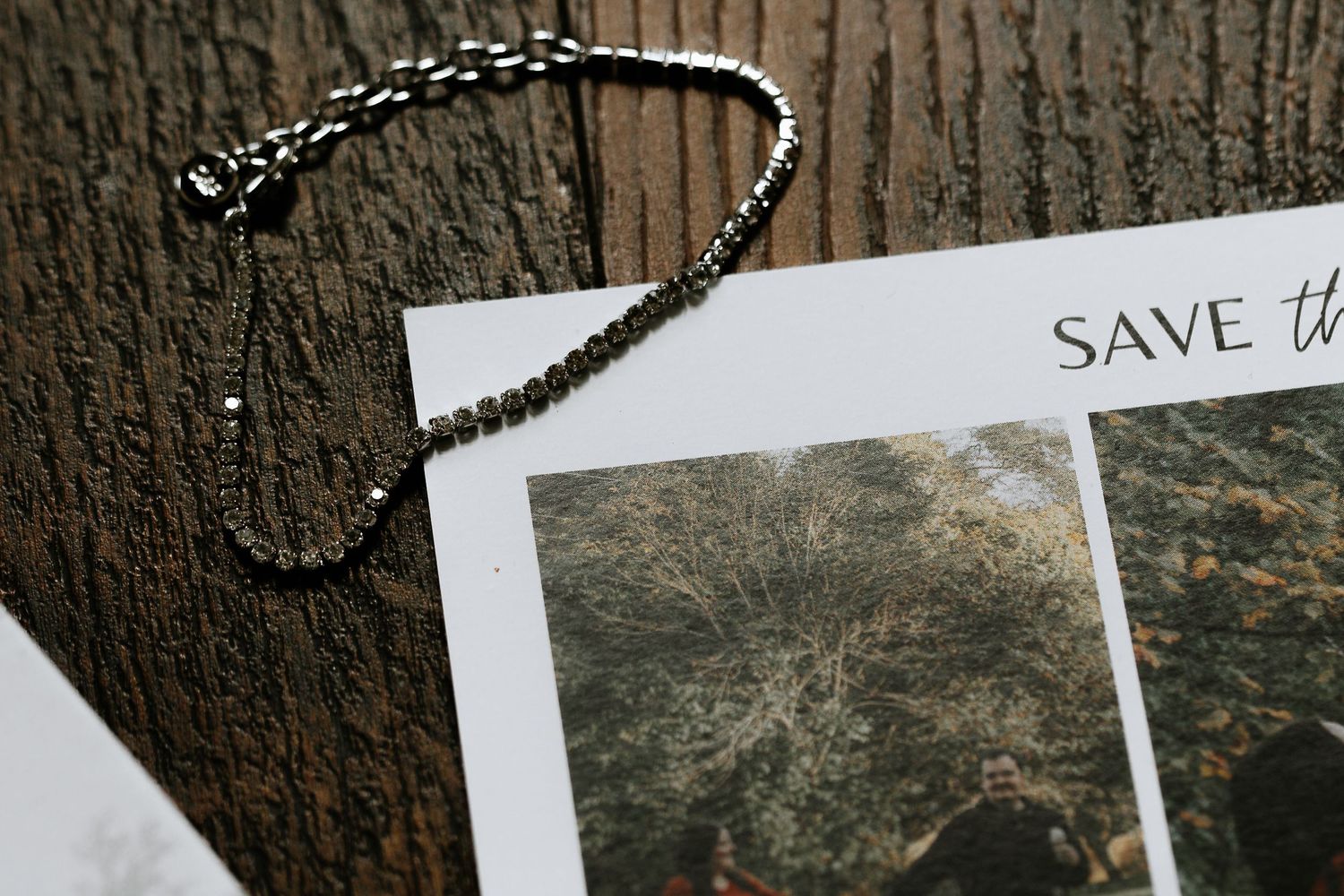 Silver chain bracelet draped over a save the date card with forest photo on rustic wood surface.