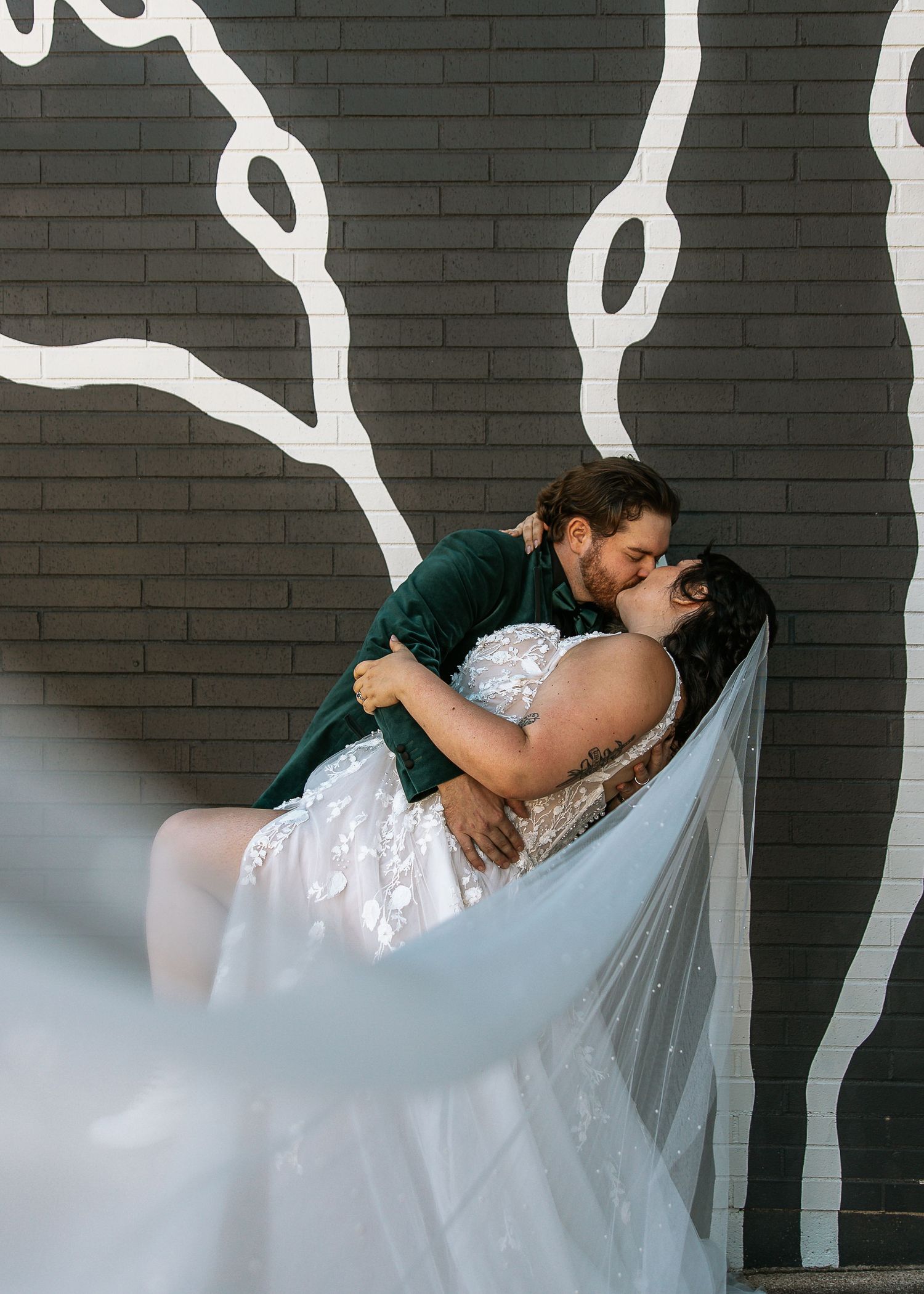 Groom in green velvet suit dips bride in lace gown for kiss against dark brick mural wall.