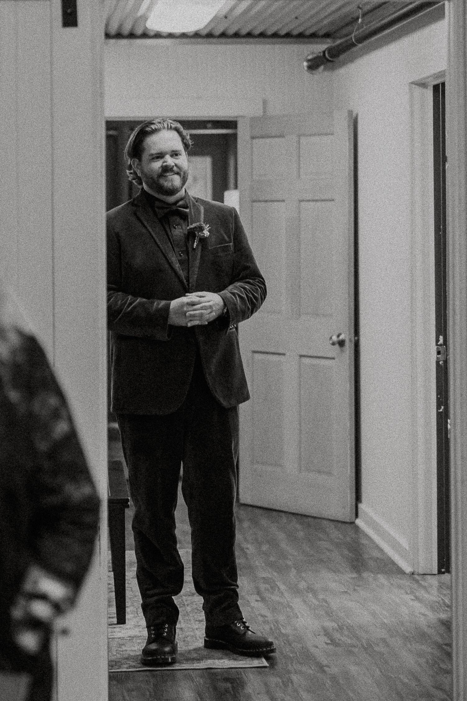Black and white portrait of groom in dark suit with boutonniere smiling in doorway before ceremony.