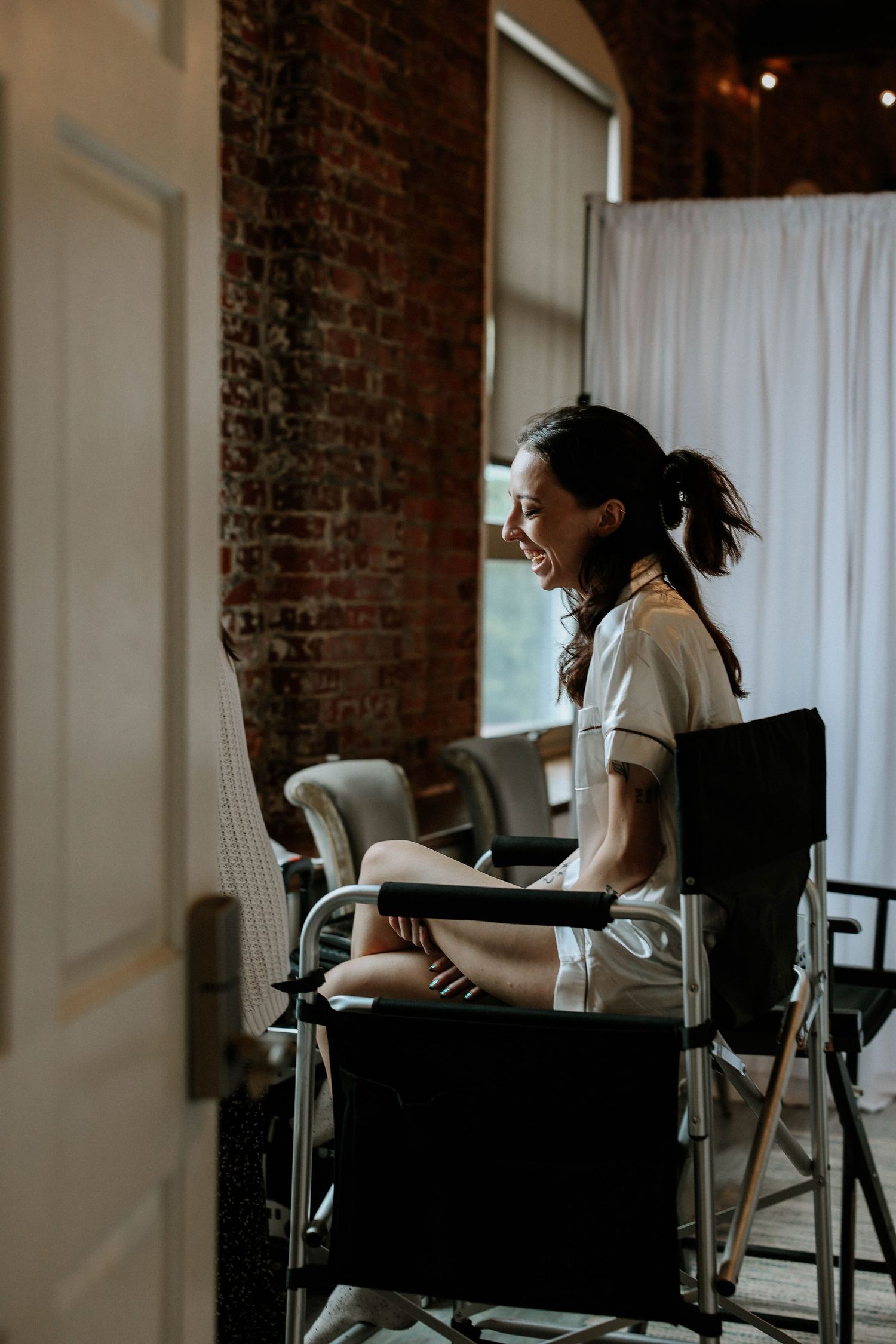 A smiling brunette woman sits in a director's chair in a rustic brick-walled room with white curtains.