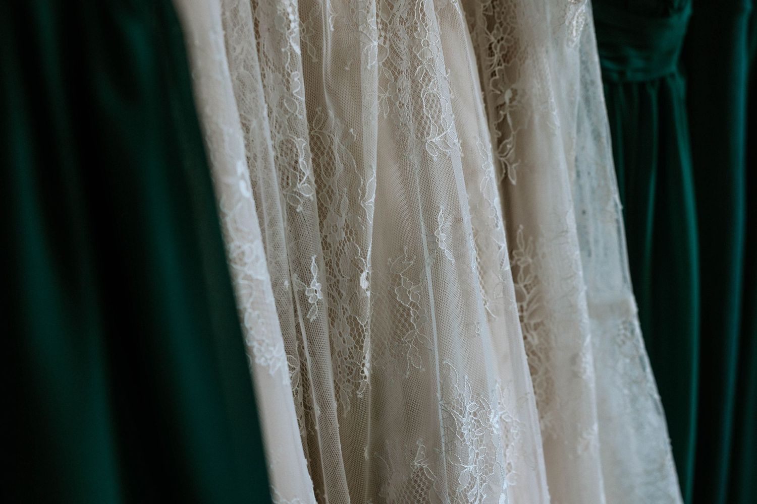 Close-up of white lace wedding dress hanging beside emerald green velvet bridesmaid dresses.