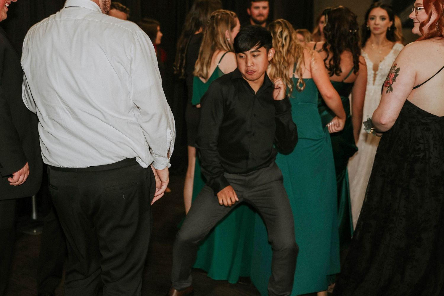 Young man dancing energetically at a formal event, surrounded by guests in elegant green and black attire.