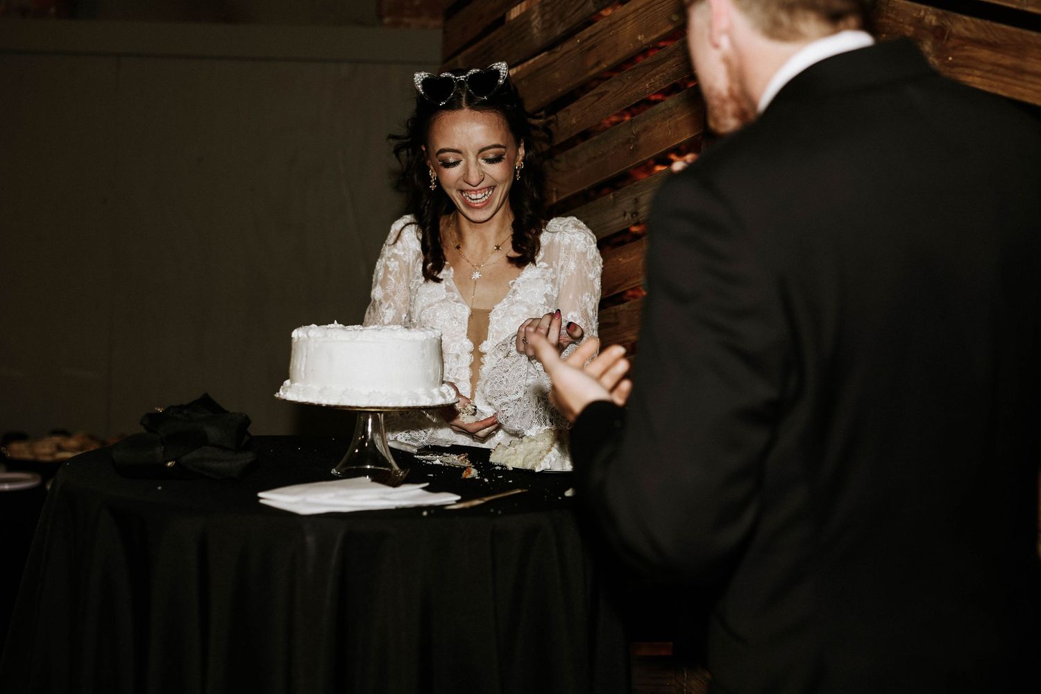 Bride in lace wedding dress smiling while cutting white wedding cake at elegant reception venue.