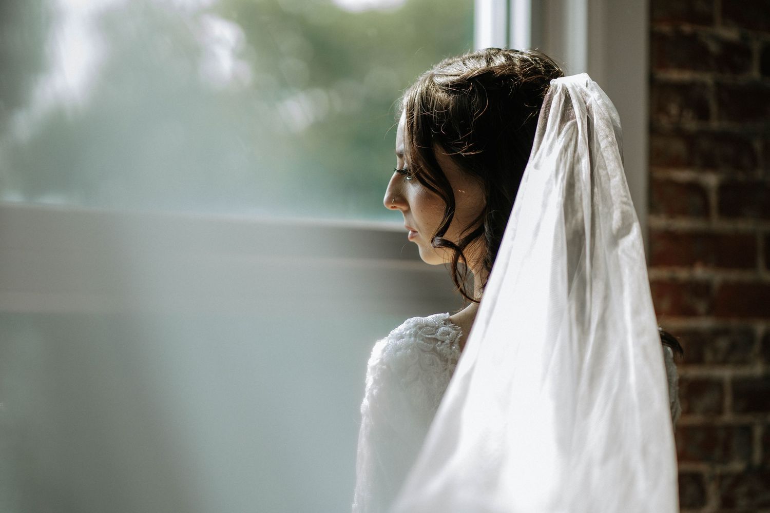 Bride in white veil gazing thoughtfully out a window, soft natural light illuminating her elegant updo and lace gown.