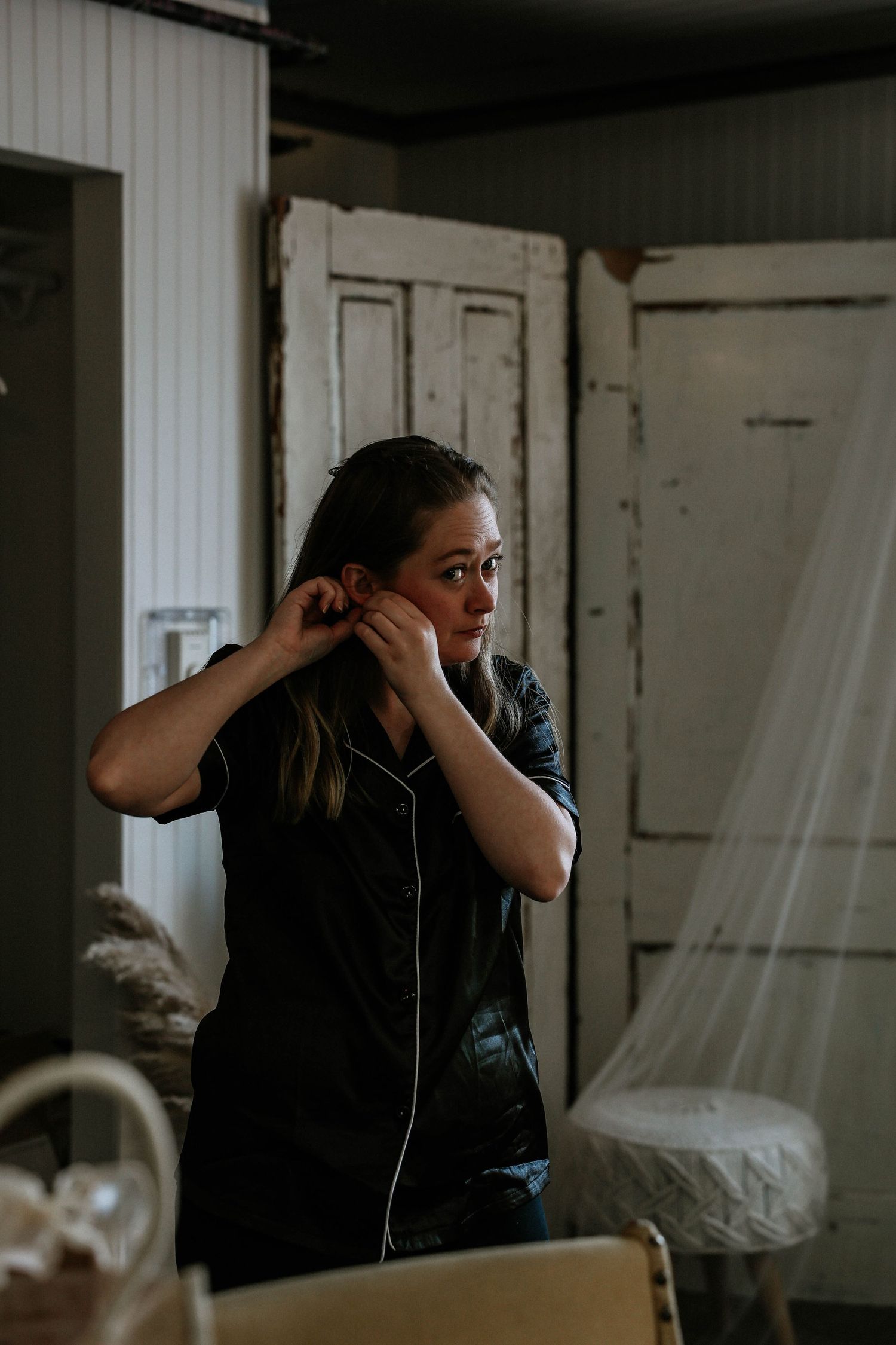 Young woman in black satin pajamas putting on earrings in a rustic vintage dressing room.