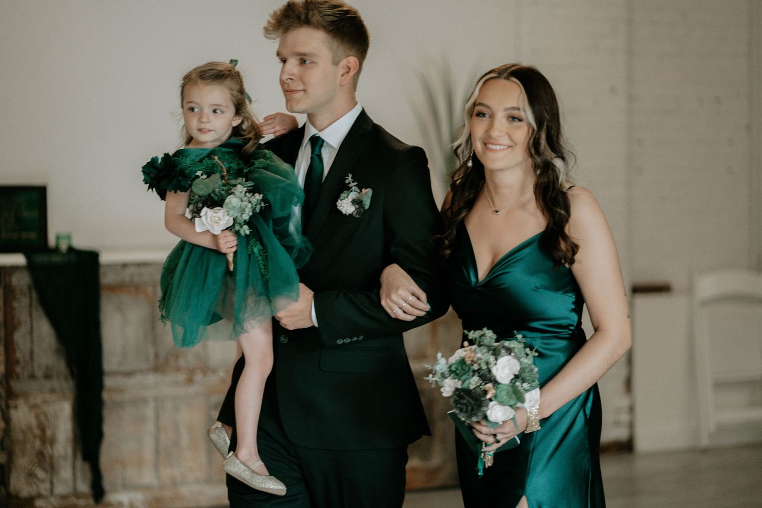Man in tuxedo holds flower girl in green tulle dress beside bridesmaid in emerald gown holding white bouquet.