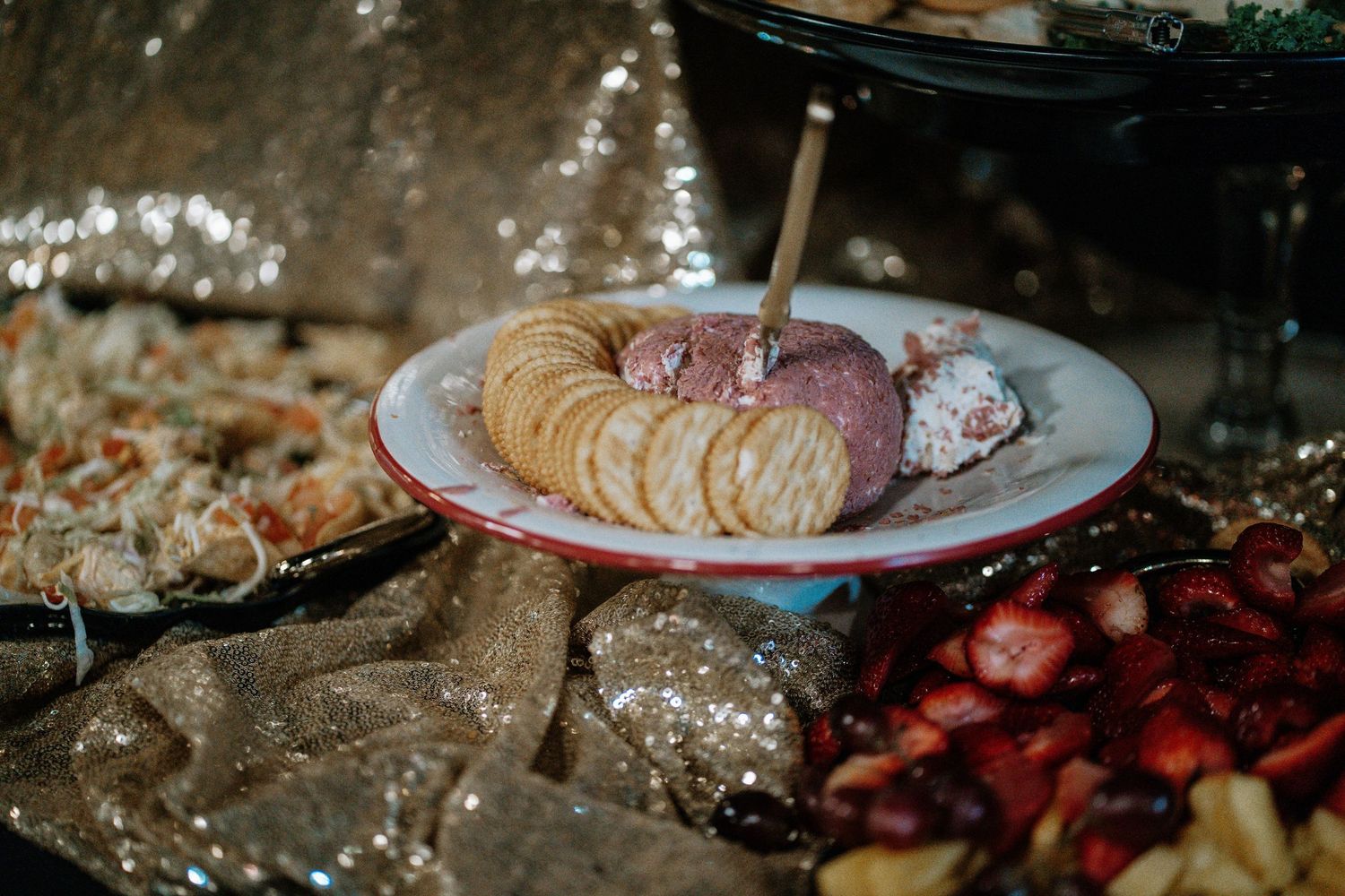 A festive party spread featuring a cheese ball with crackers, fresh strawberries, and assorted appetizers on a glittery tablecloth.