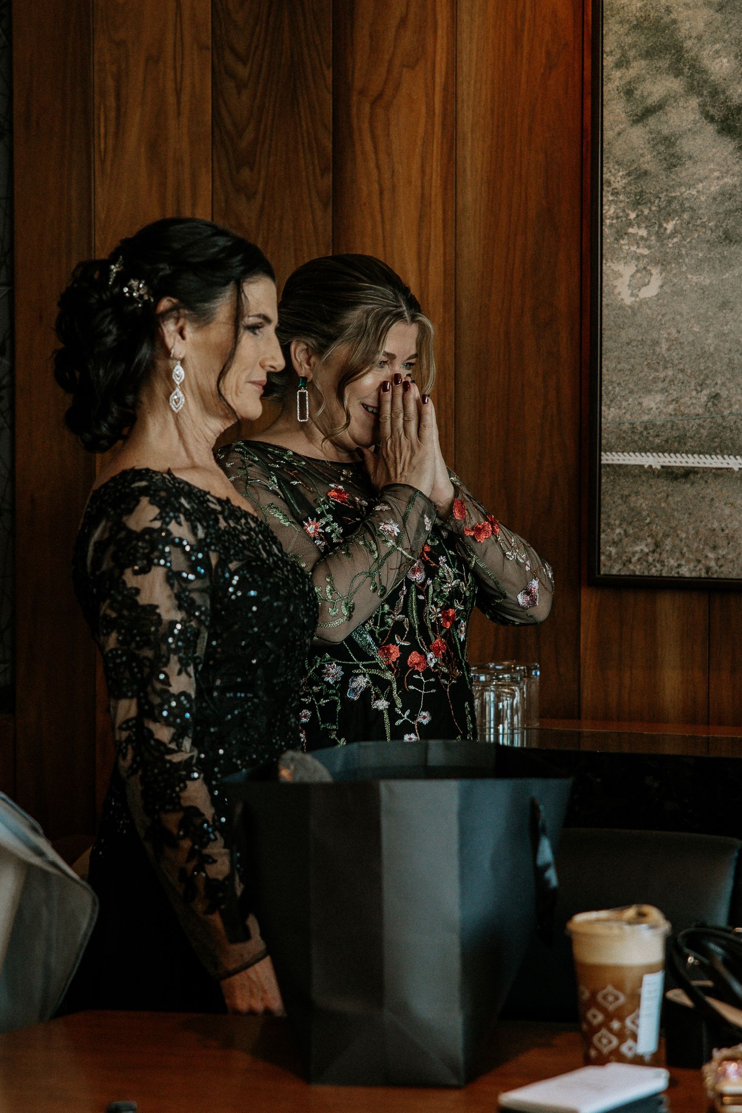 Two women in elegant black floral dresses share an emotional moment, one covering her mouth in surprise at a wedding.
