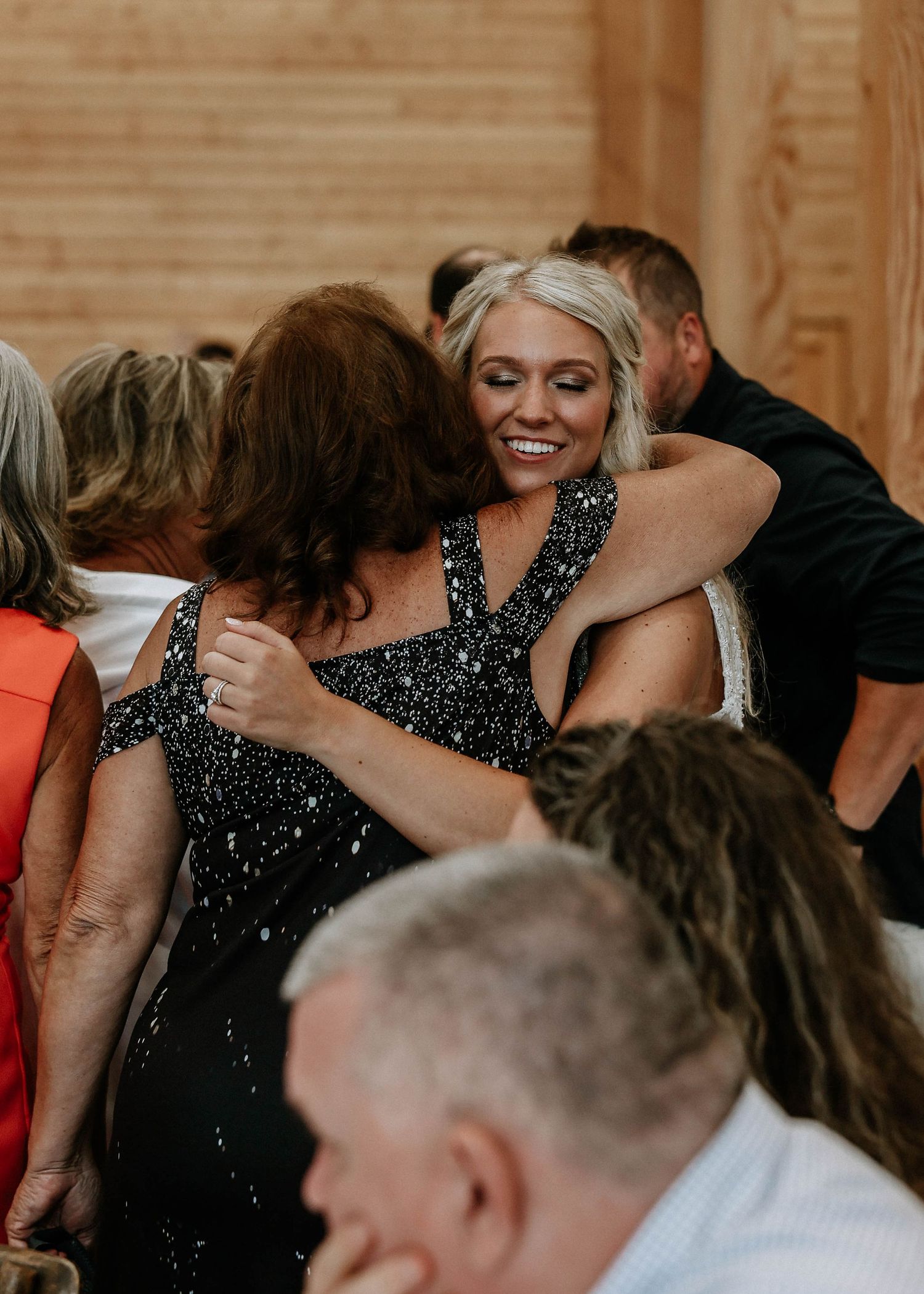 Two women in elegant black dresses share a warm hug at a formal event inside a wooden venue.