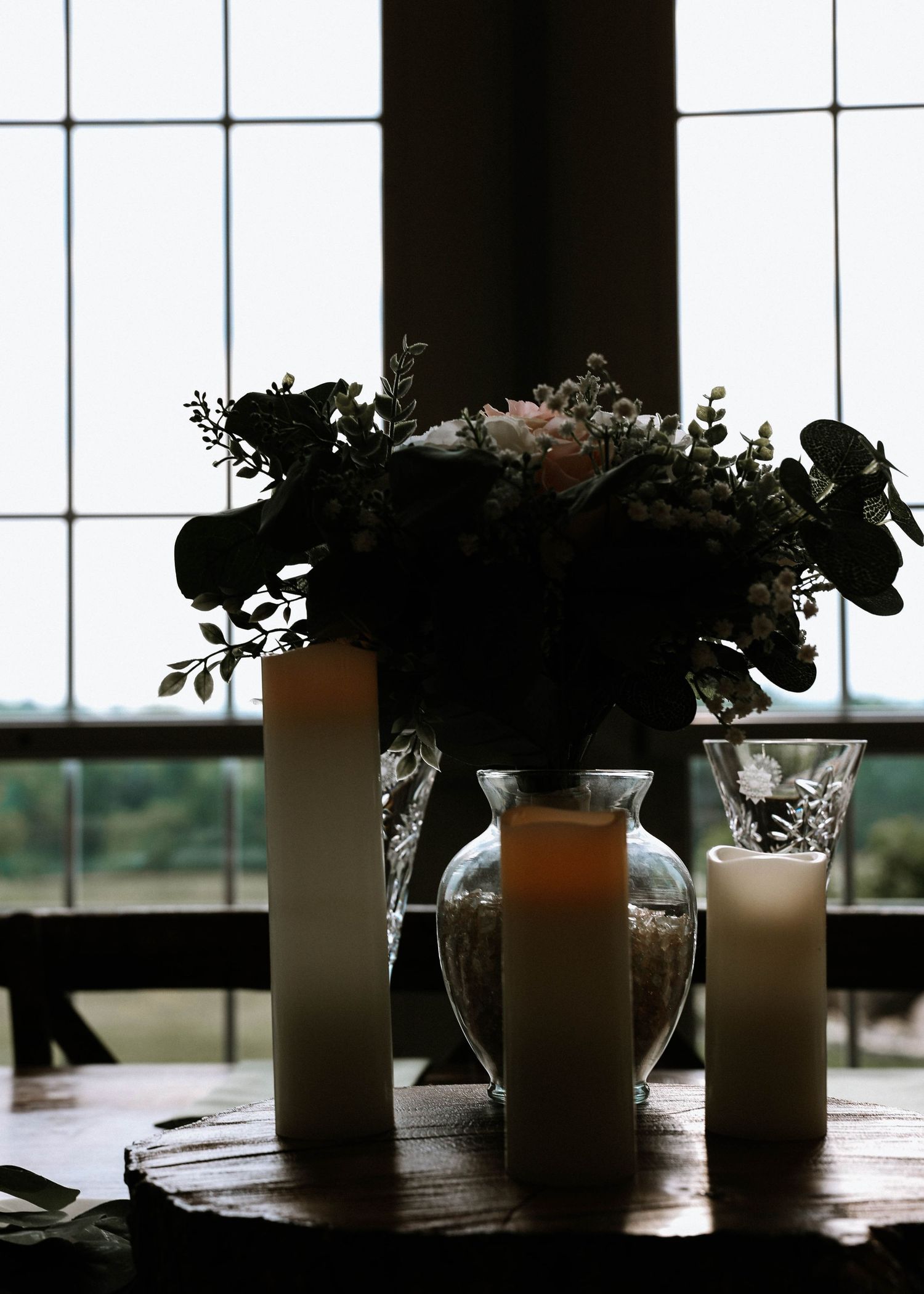 Elegant wedding centerpiece with crystal vase of pink flowers, greenery, and white pillar candles on rustic table.