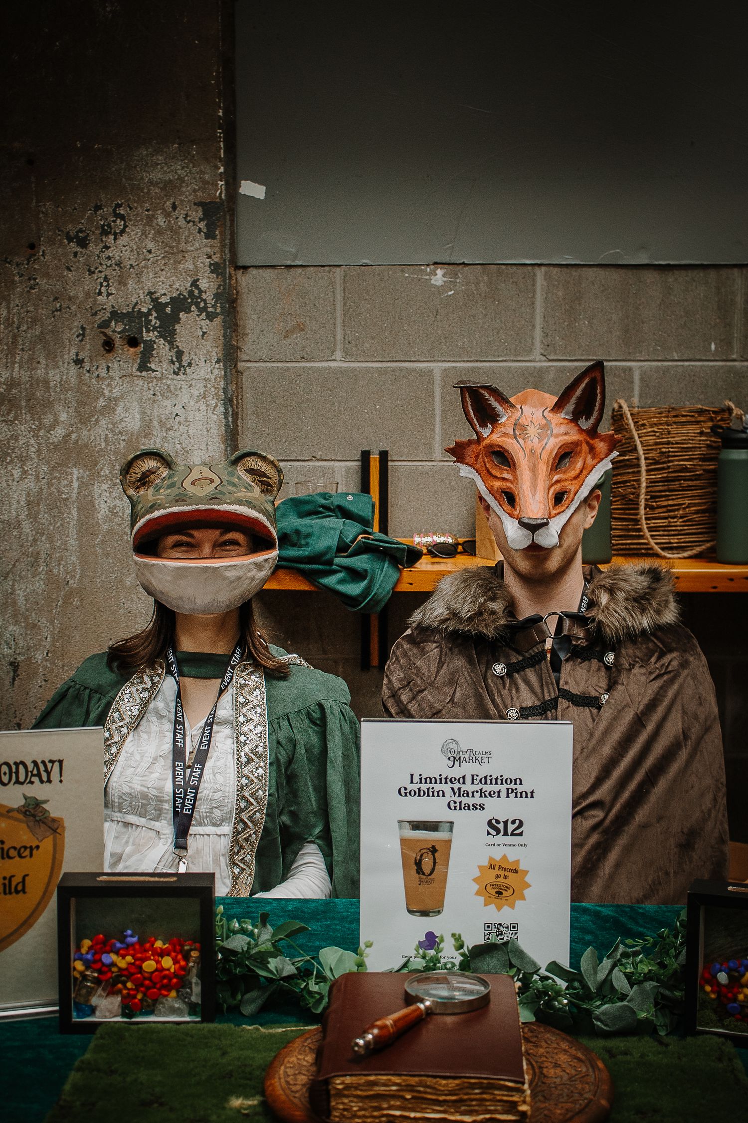 Two people wearing animal masks at a Goblin Market booth selling limited edition pint glasses for $12.