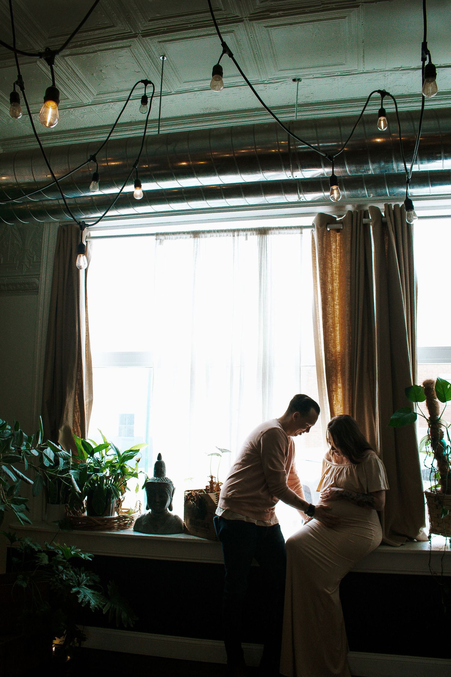 Couple sharing intimate moment by window with hanging Edison bulbs in Cincinnati, Ohio.