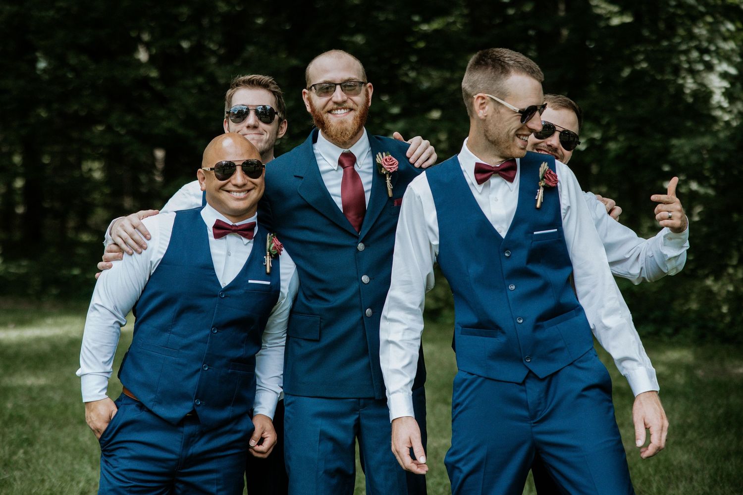 Four groomsmen in navy blue vests and bow ties pose playfully wearing sunglasses in a wooded outdoor setting.