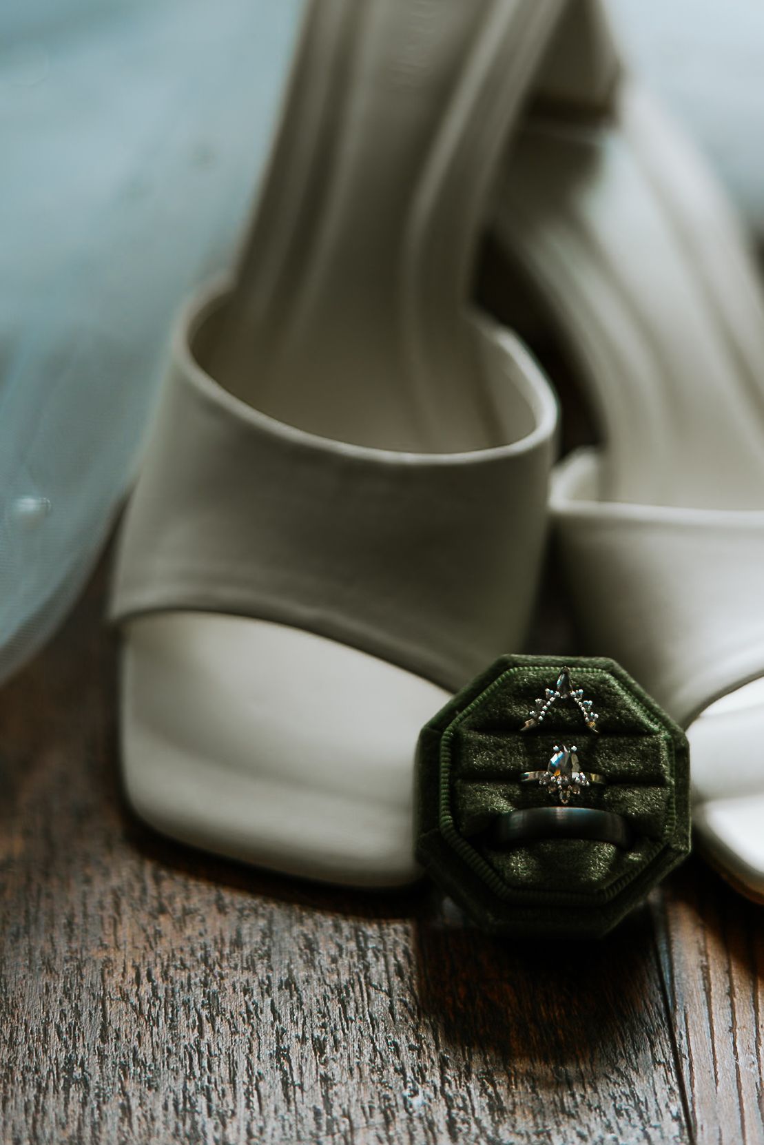 Wedding rings displayed in a green velvet octagonal ring box beside white bridal heels on wood.