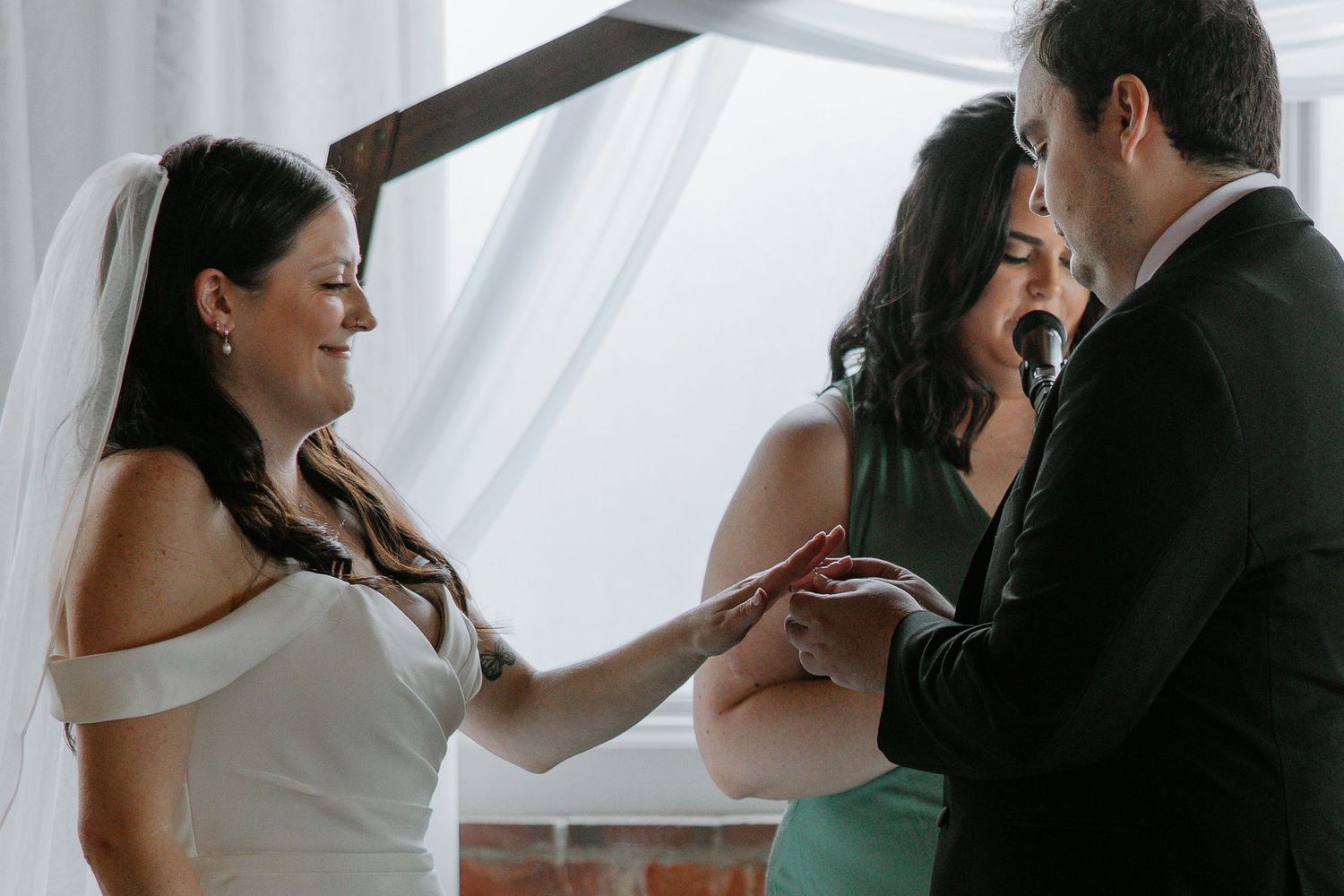 Bride placing wedding ring on grooms finger during indoor wedding ceremony.