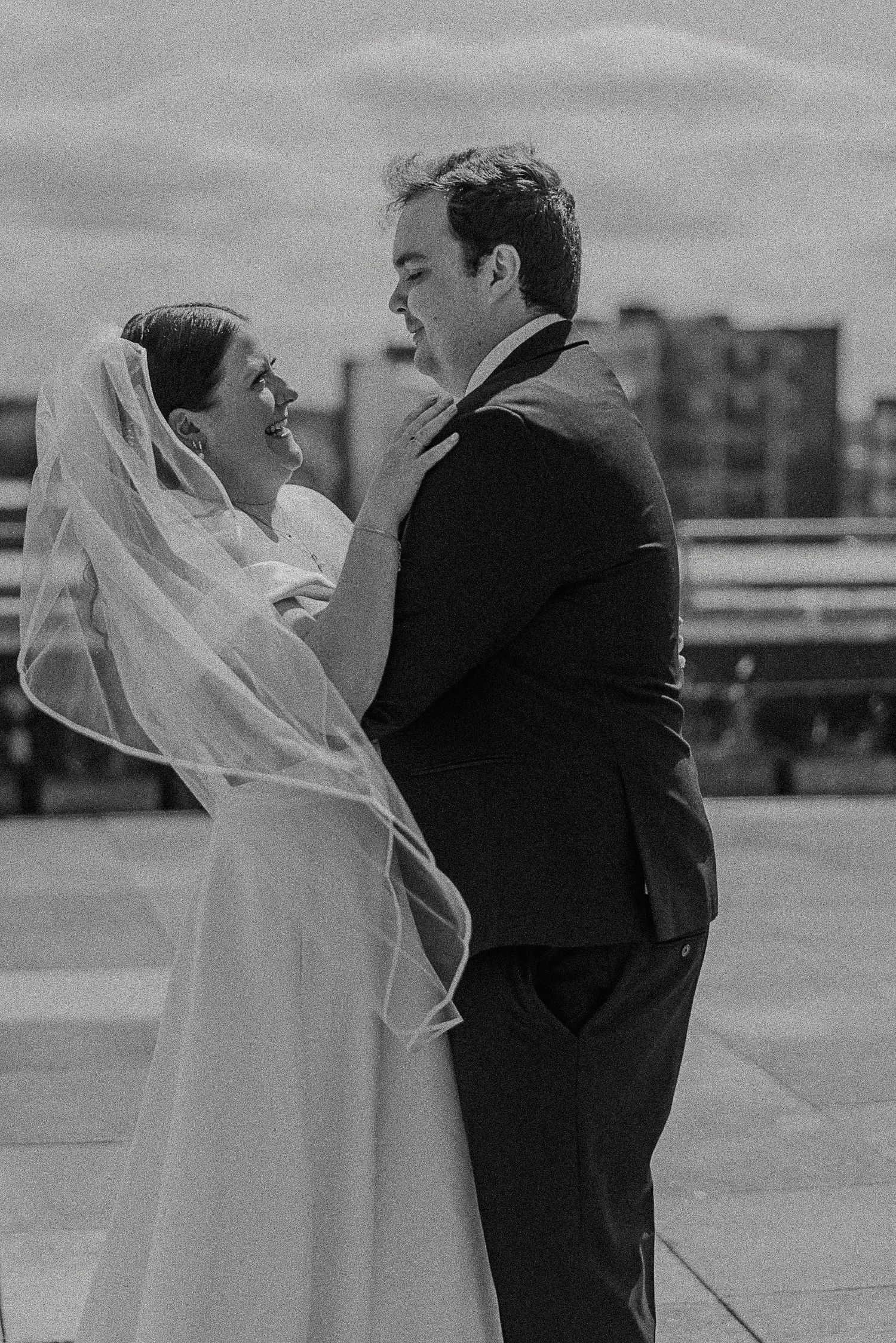 Black and white photo of smiling bride and groom dancing on rooftop, bride in elegant veil.