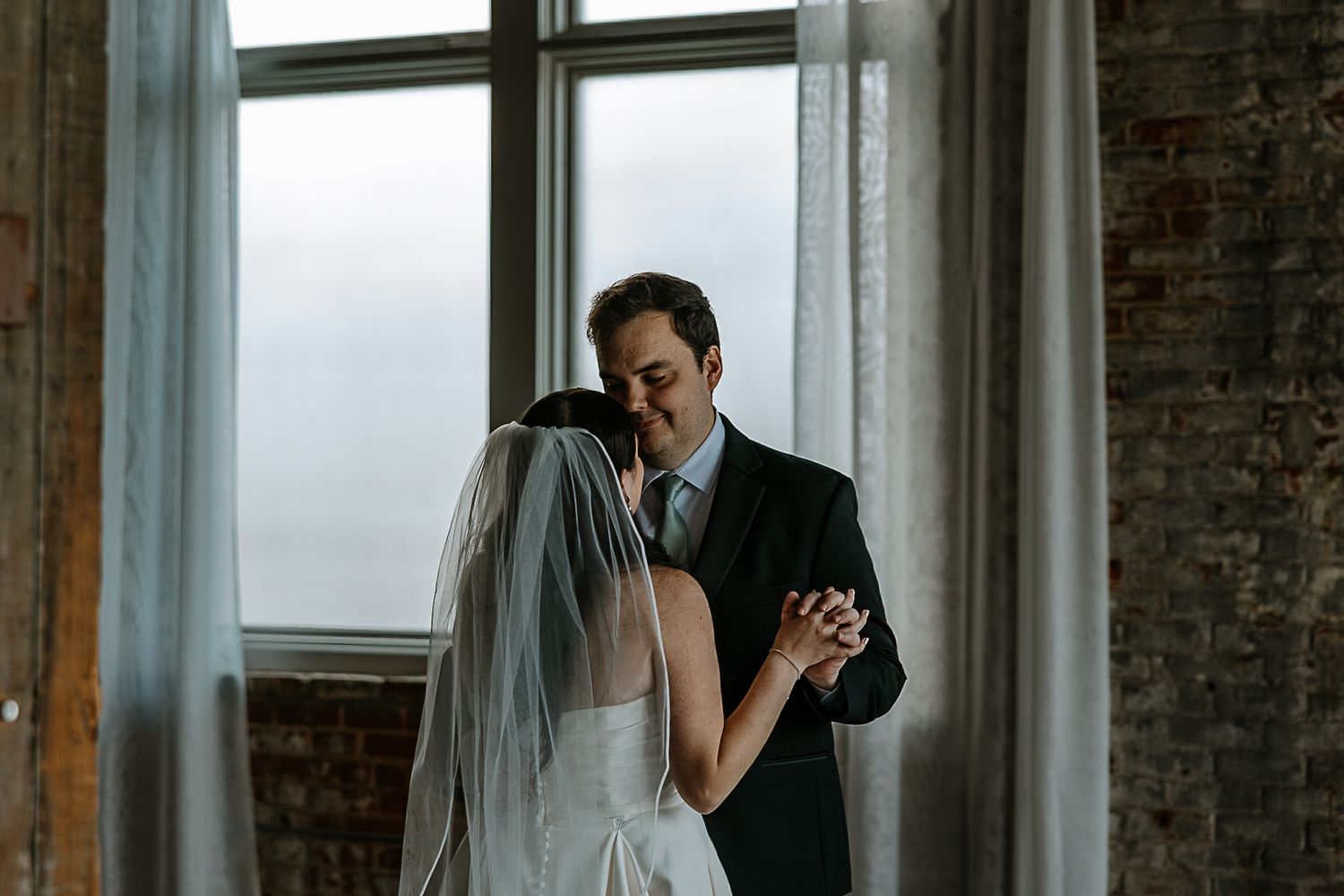 Bride and groom share an intimate first dance moment by large windows in a rustic brick venue.