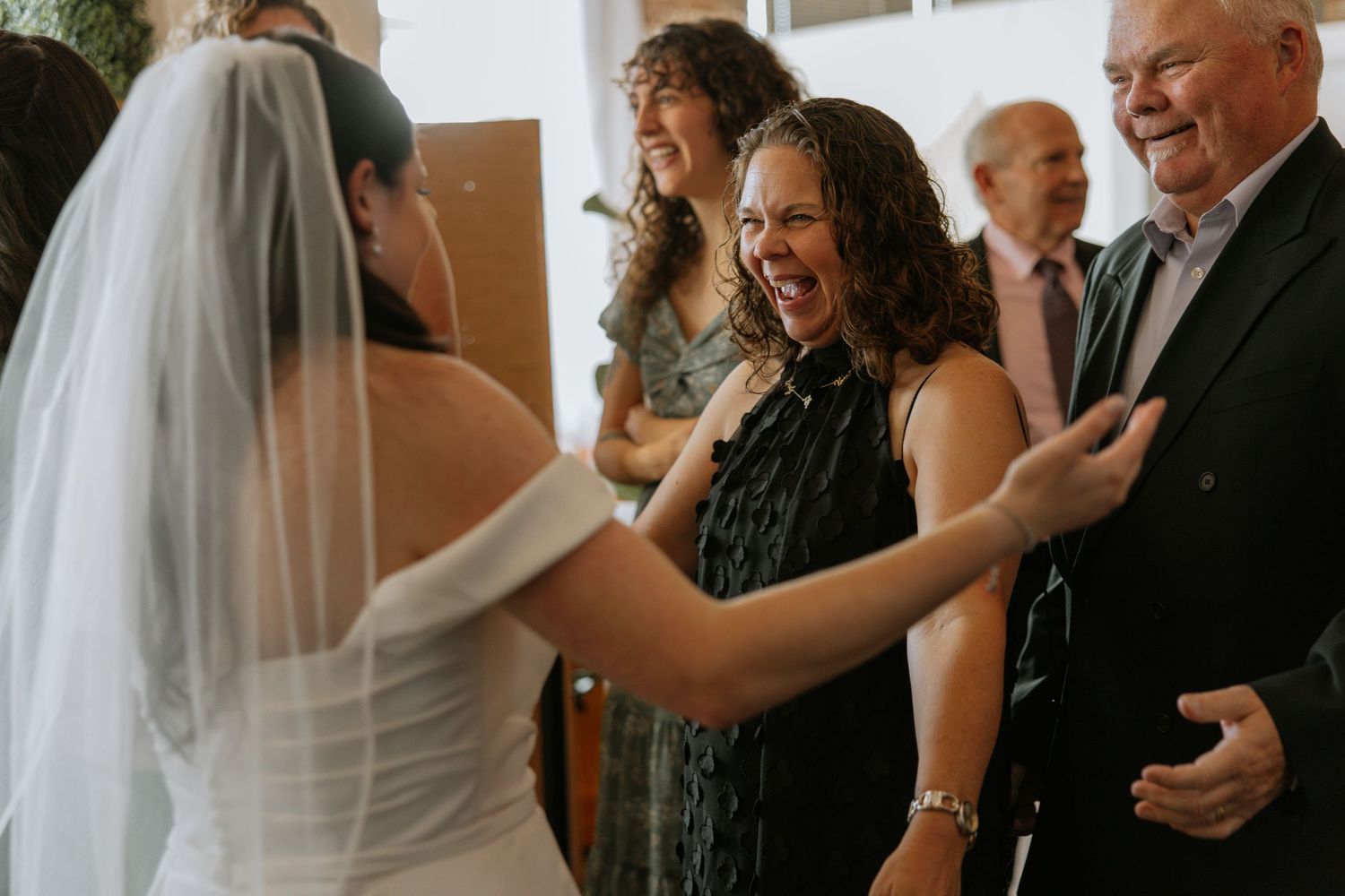 Bride in white veil greeted by laughing female guest and older man at wedding reception.